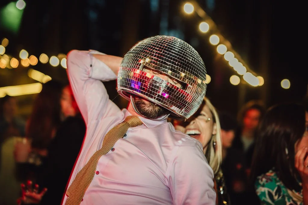 A person wearing a disco ball helmet and light pink shirt enjoying a party with friends under string lights.