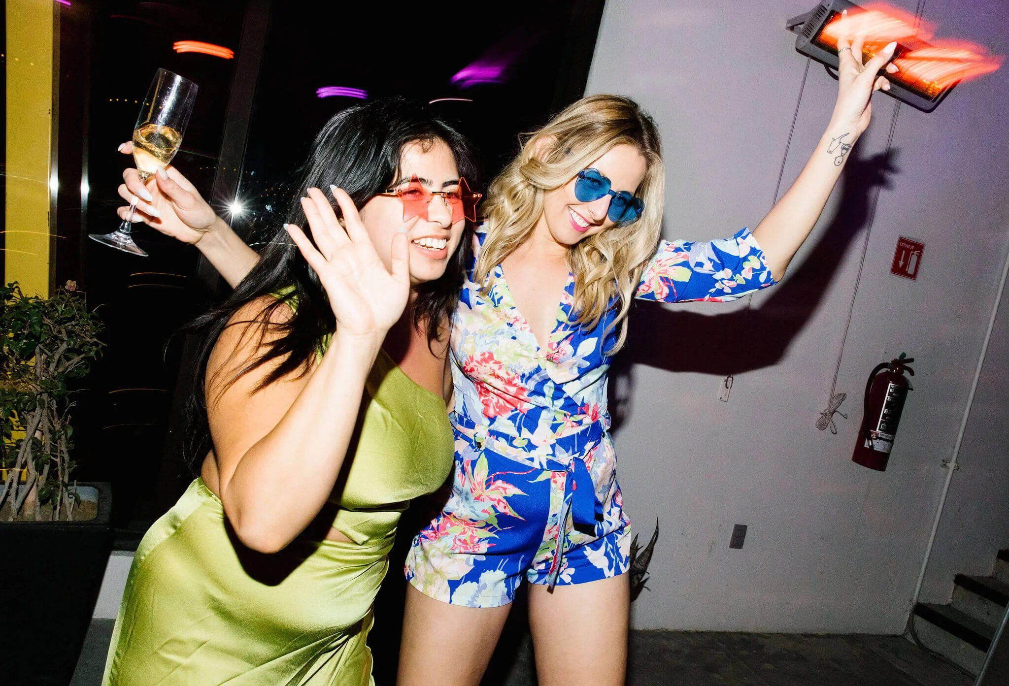 Two women celebrating at a party, one wearing red star-shaped sunglasses with a champagne glass, and the other wearing blue sunglasses with colorful floral pajamas, dancing with raised arms.