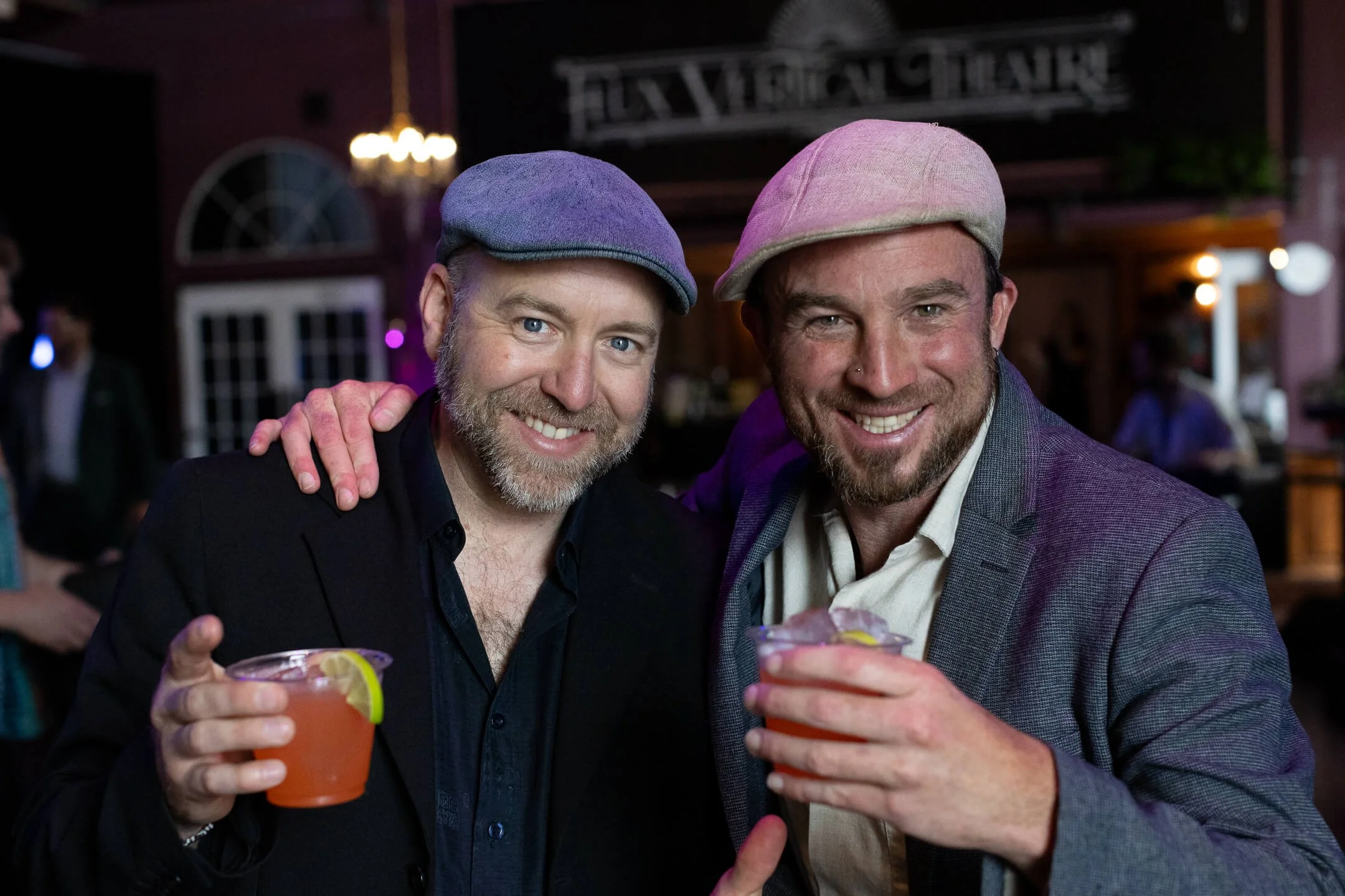 Two men smiling at a bar, holding cocktails garnished with lemon slices, surrounded by a lively atmosphere with other patrons and a bar sign in the background.