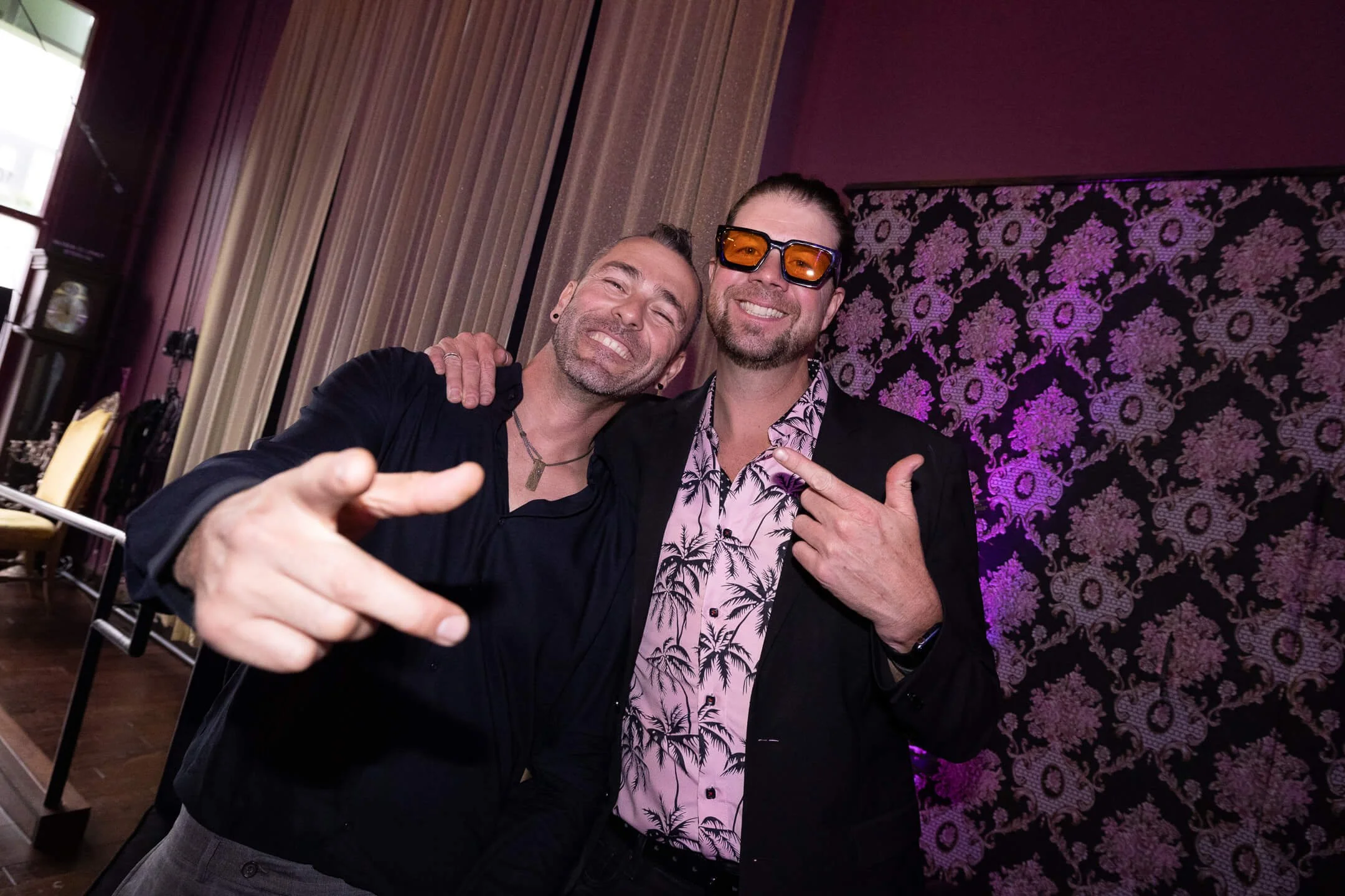 Two men smiling and posing together at a social event, one wearing sunglasses and a Hawaiian shirt, the other wearing a black jacket, indoors with decorative wallpaper and curtains in the background.
