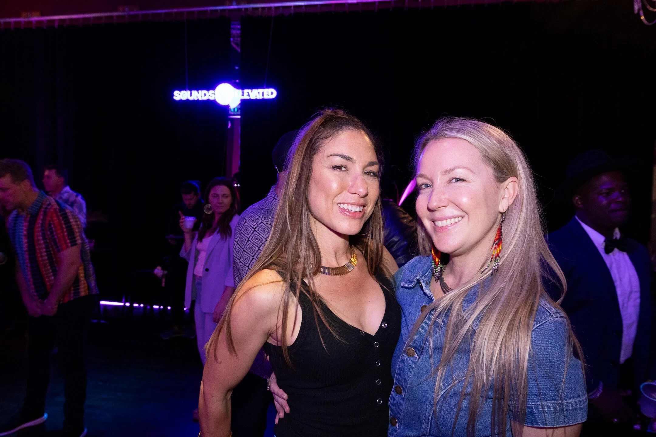 Two smiling women posing together at a nightclub or party, with a sign in the background that reads 'SOUNDS ELEVATED' and other people in the background.