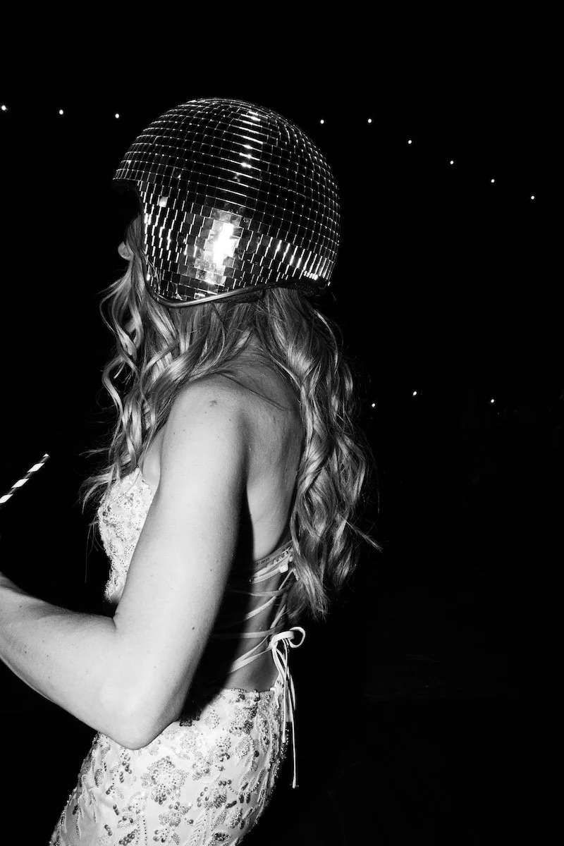 A woman with wavy hair wearing a disco ball helmet and a sleeveless, lace dress in a dark setting with small lights in the background.