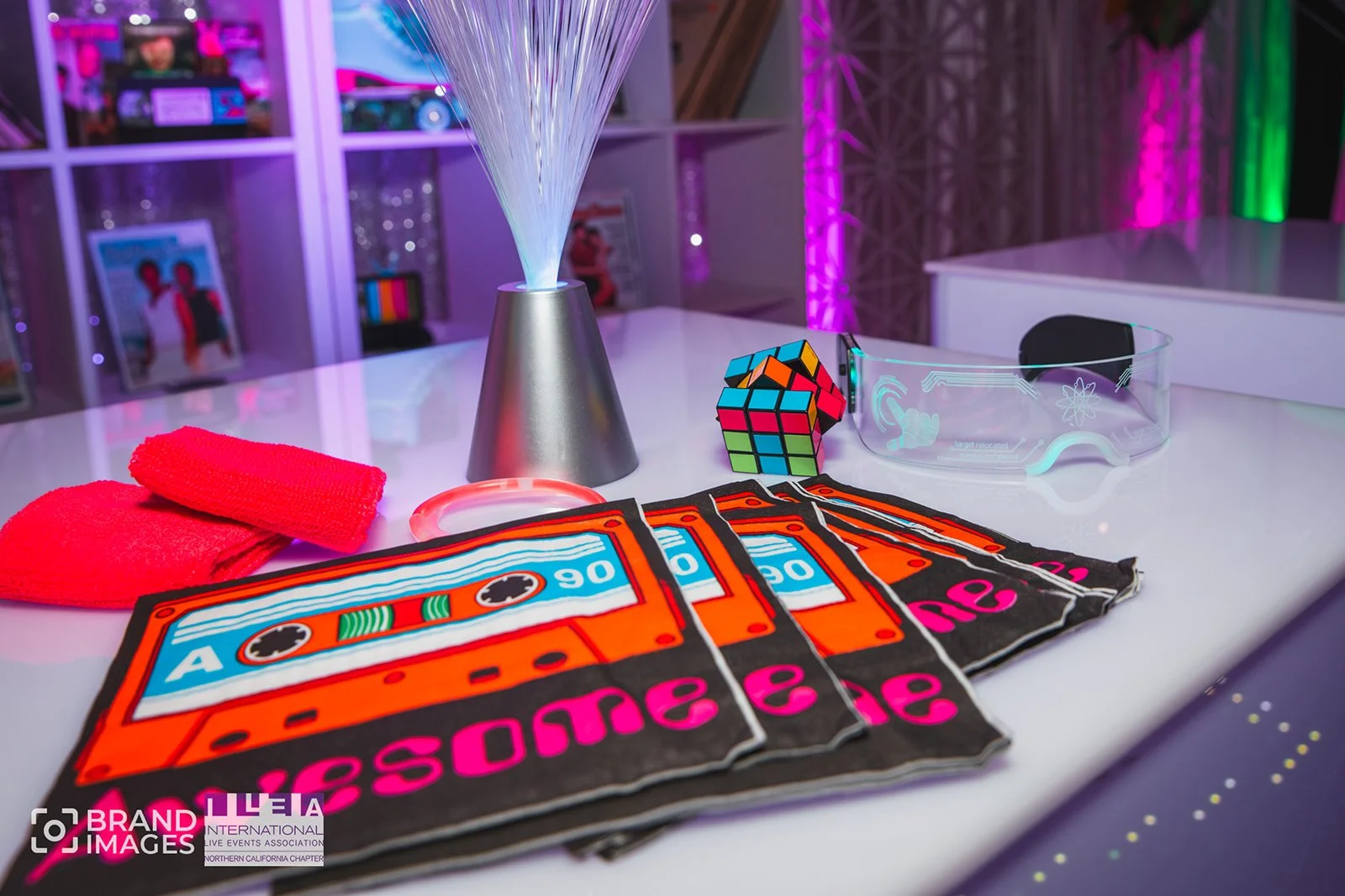Colorful glow-in-the-dark items on a table including candy cane shaped tags spelling 'awesome', glow glasses, a Rubik's cube, a fiber optic lamp, red tape, and a pink glow-in-the-dark bracelet, with a neon-lit background.