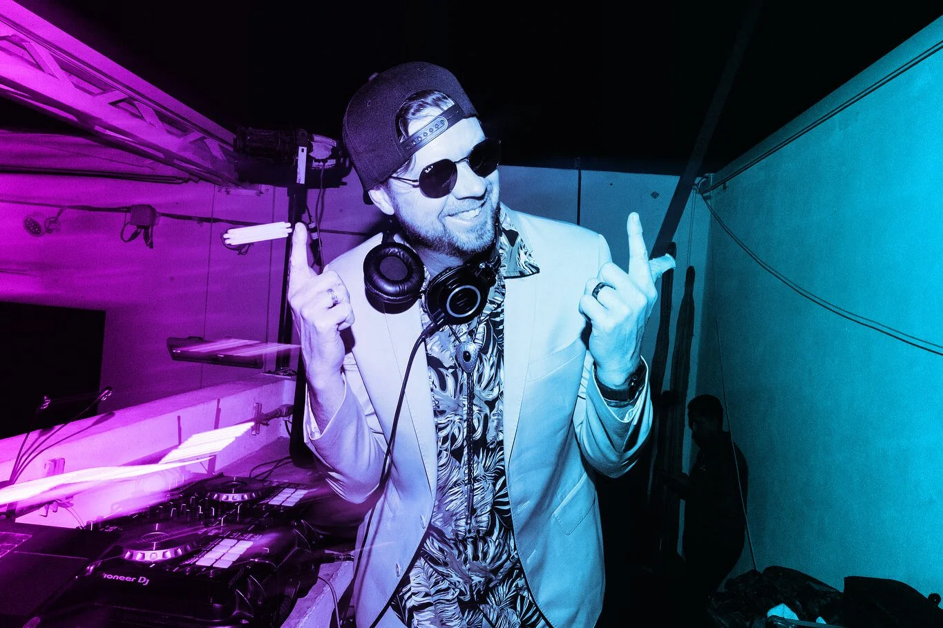 A man with a beard in a tropical shirt, blazer, sunglasses, and a backwards cap, smiling and pointing with both hands at a DJ booth with colorful lighting.