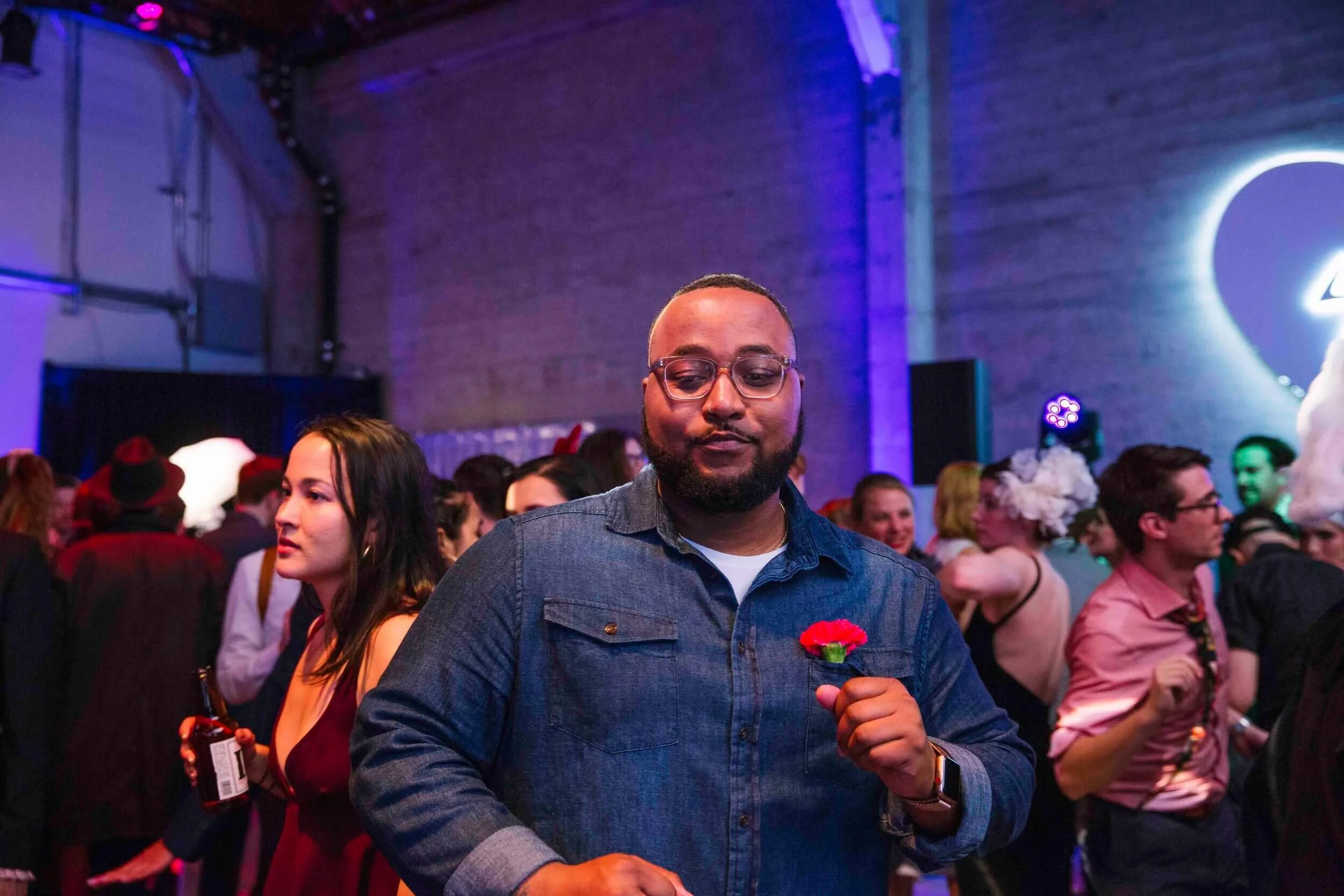 A man holding a red flower at a crowded party or event with colorful lighting and a brick wall in the background.