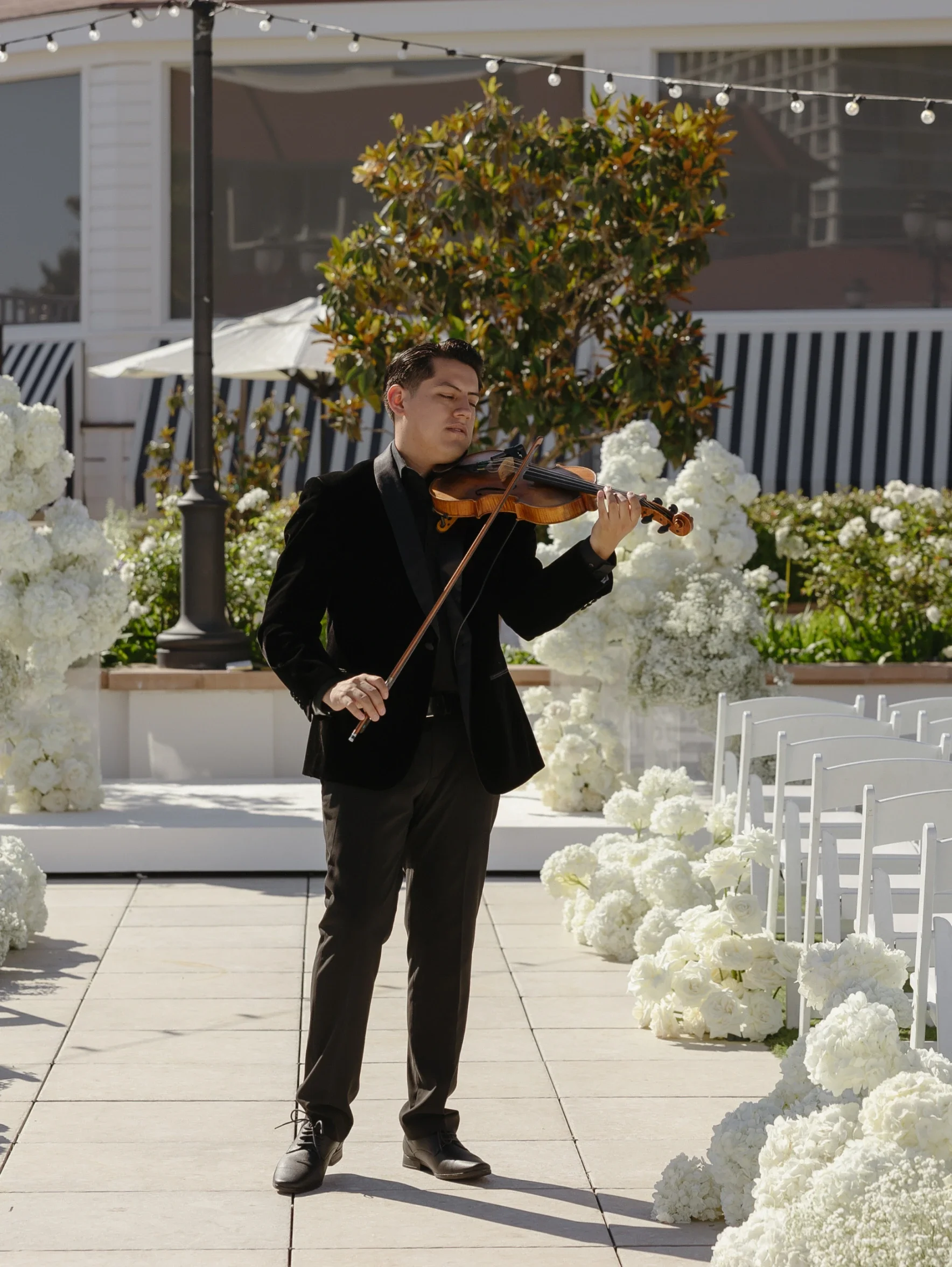 A man dressed in a black suit playing the violin outdoors, surrounded by white flowers and chairs, with a tree and building in the background, likely with wedding dj at an outdoor event.