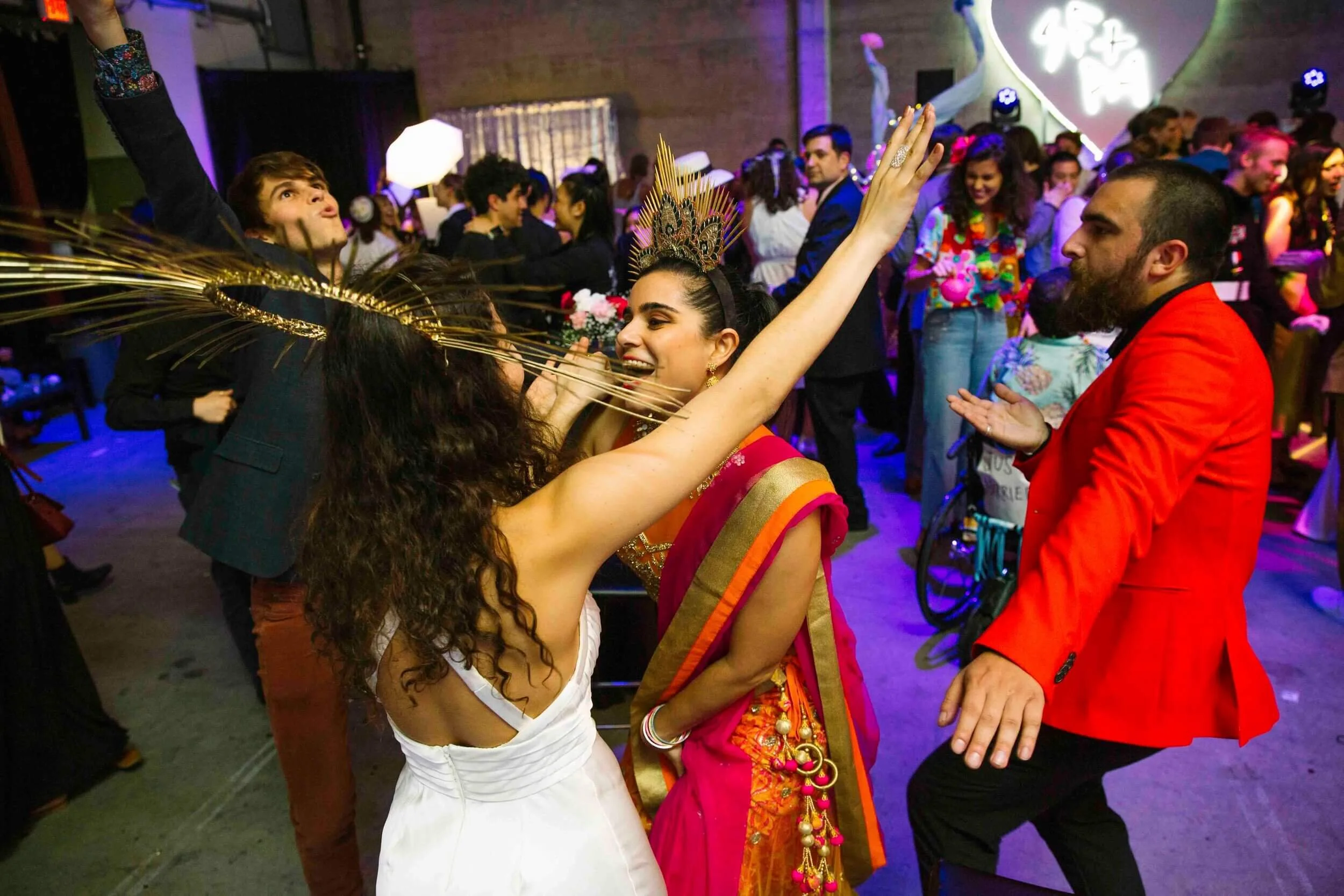 People dancing and celebrating at a party with colorful lighting. A woman dressed in traditional Indian attire, wearing a crown, is smiling, while another woman in a white dress with gold accessories is dancing with her arms raised. A man in a red ja