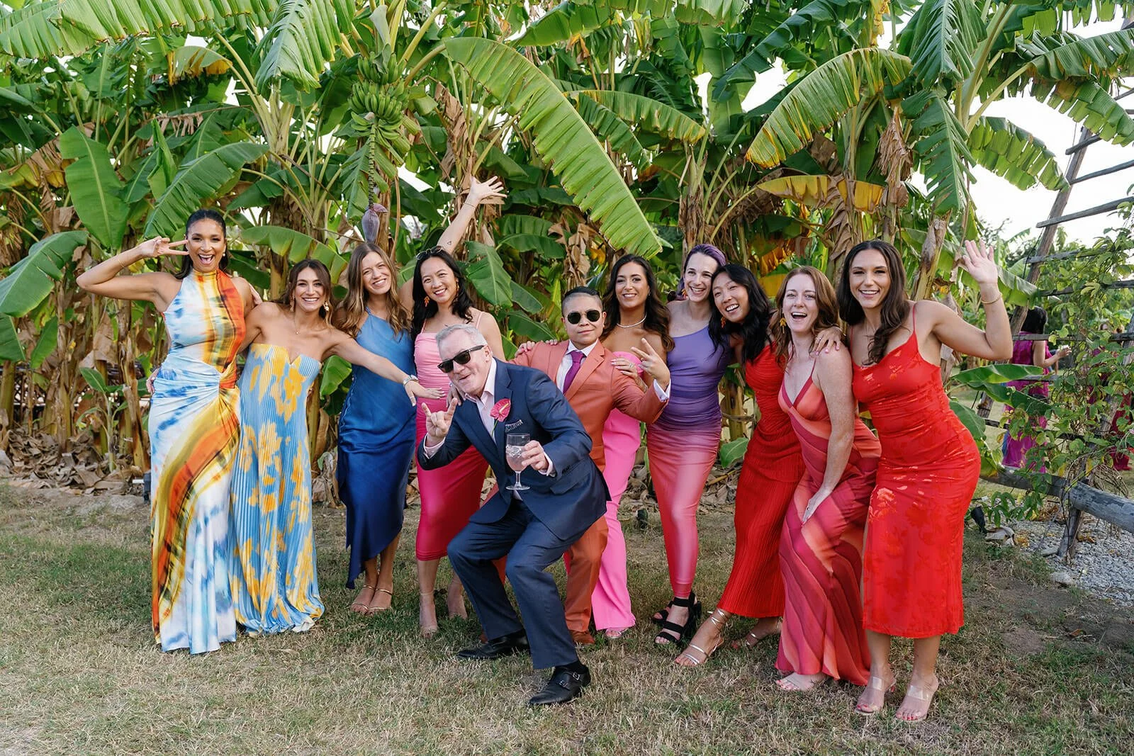 A group of eleven people, including men and women, dressed in colorful party attire, standing outdoors in front of banana plants, enjoying a celebration or gathering and posing for a photo.