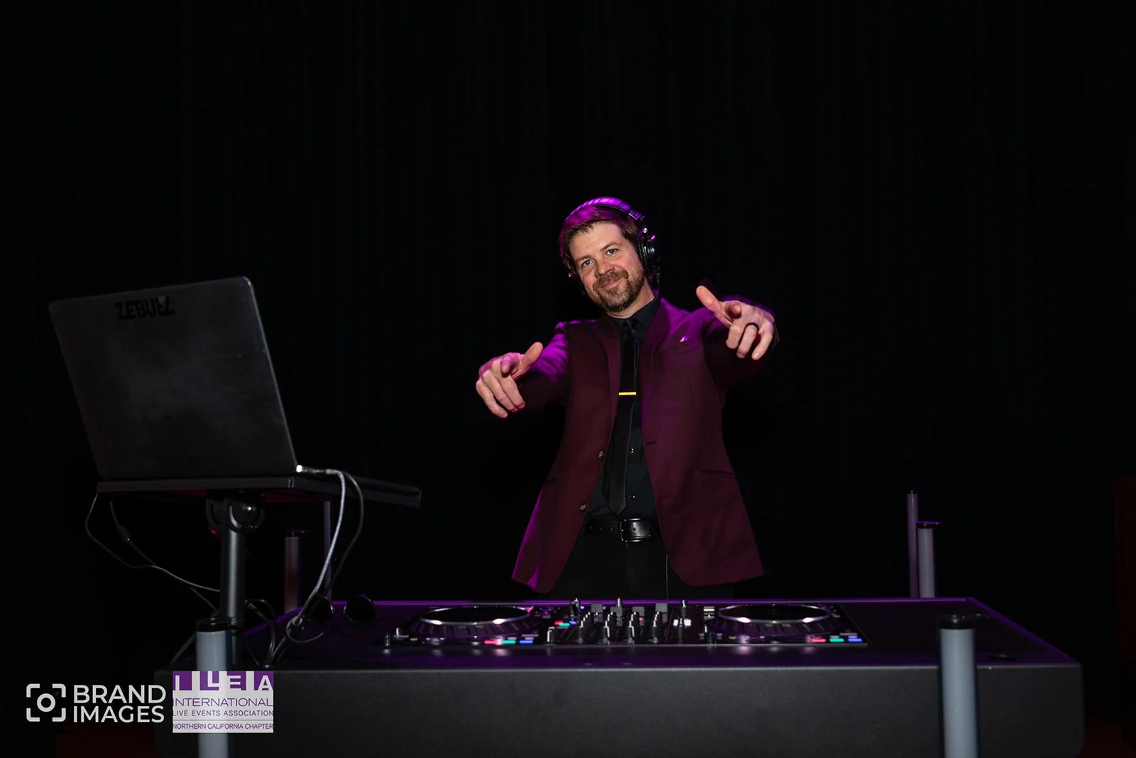 DJ wearing headphones and a maroon blazer, standing behind a turntable at a dark event space, pointing towards the camera with both hands.