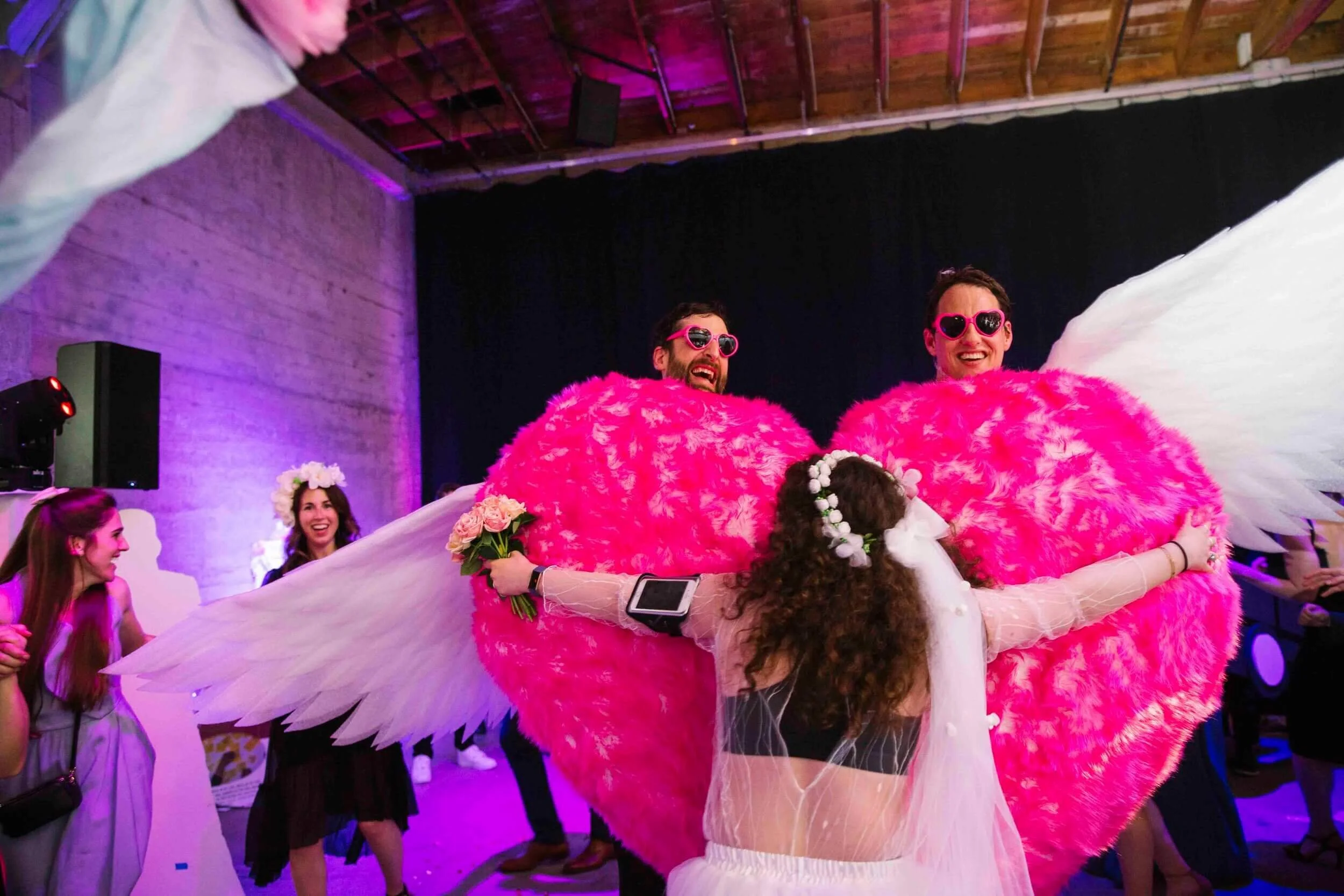 People celebrating at a party, wearing costumes with pink fluffy angel wings and heart-shaped sunglasses. A woman dressed as an angel with a floral crown is hugging two men dressed as angels with large pink fluffy hearts and white wings.