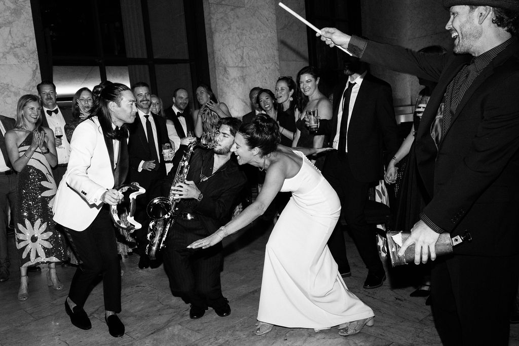 People dancing at a party with a live band, a woman in a wedding dress, and a man with a saxophone, all enjoying the music and laughter.