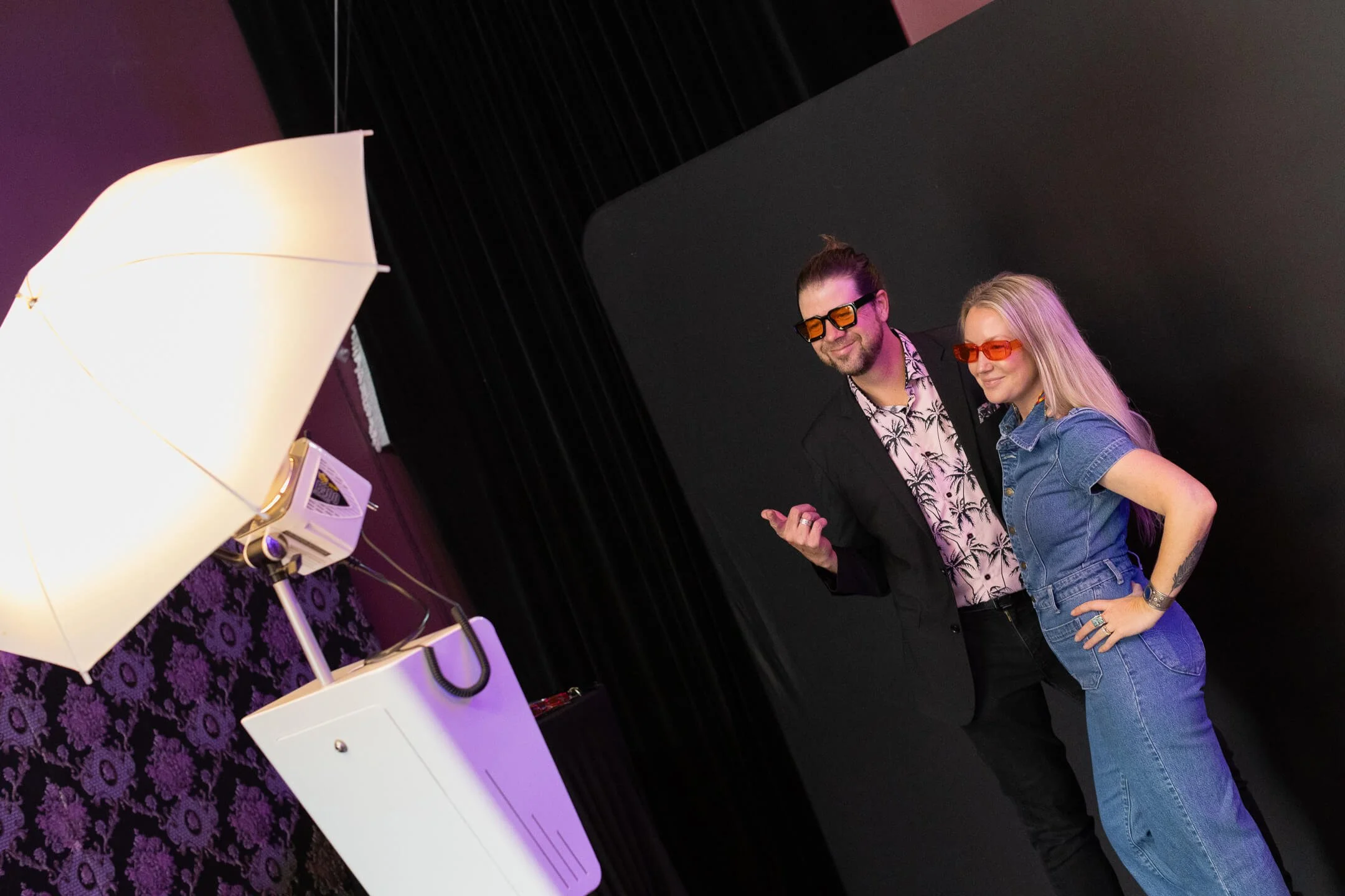 A man and woman wearing 3D glasses posing for a photo in front of a black background, with a purple patterned wall and studio lighting equipment nearby.
