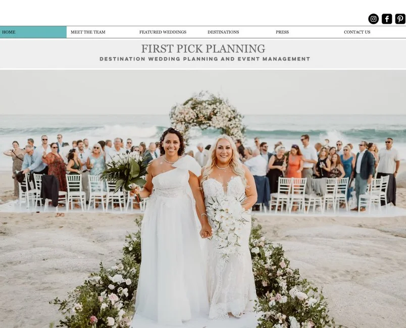 Two women in wedding dresses walking down the beach aisle at a wedding ceremony. Attendees are seated behind them, with the ocean and a floral arch in the background.