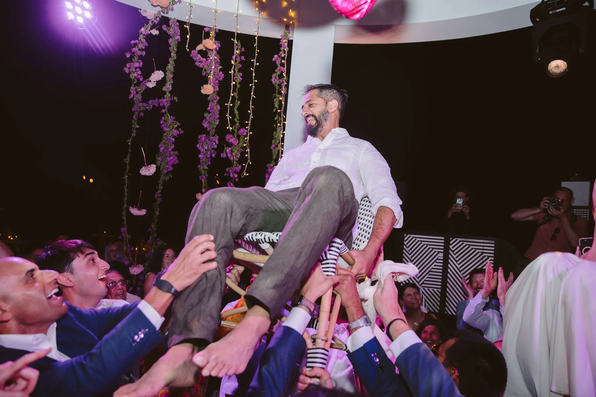 A man wearing a white shirt sits on a chair held up by a crowd of people at a celebration, with decorative hanging flowers and lights in the background. The crowd appears joyful and is supporting the man.