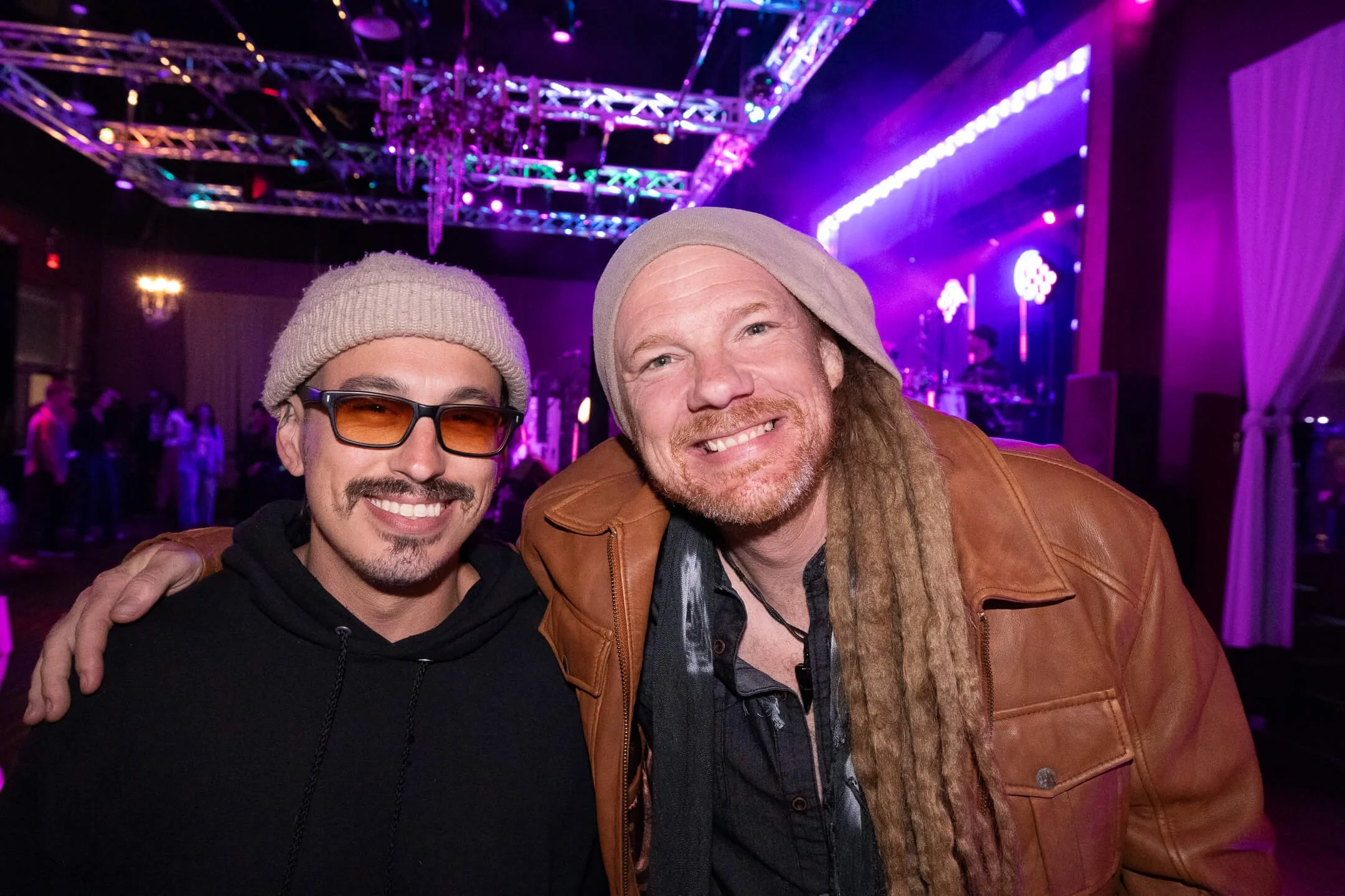Two smiling men at an indoor concert or event with purple lights, a stage, and other people in the background.