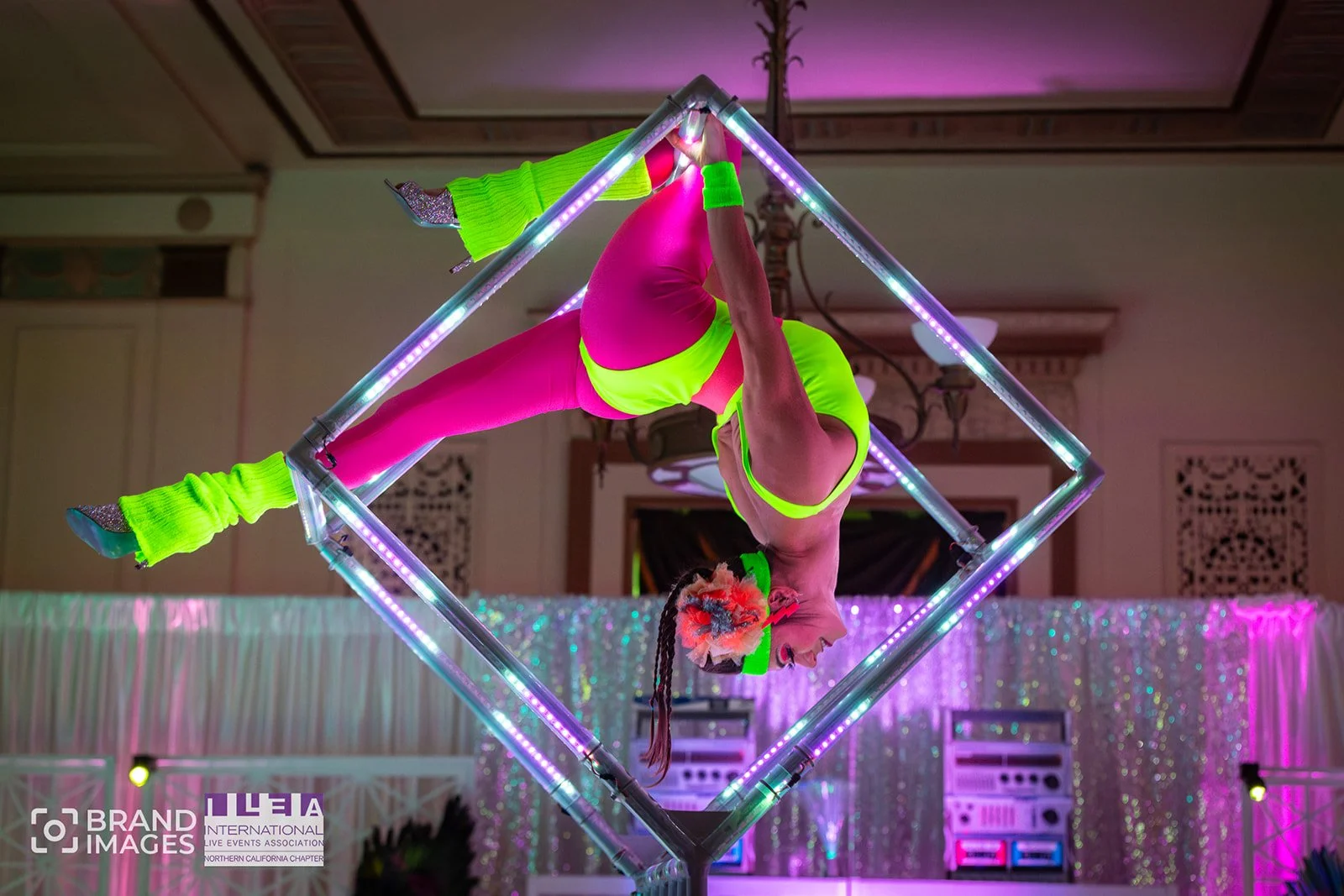 A performer in bright pink and neon green costume hanging upside down inside an illuminated square frame at an event, with a shiny backdrop and stage lights in the background.