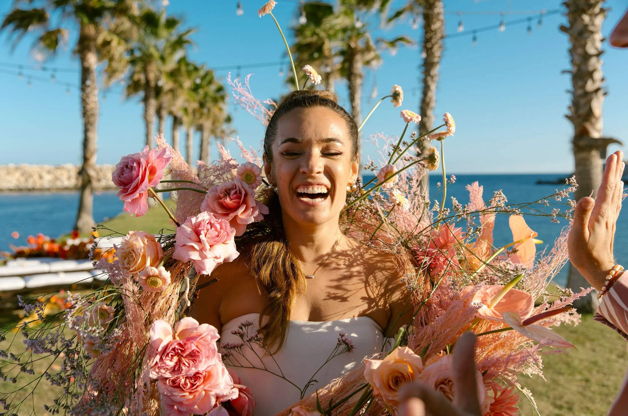Joyful woman with long hair, wearing a floral dress, surrounded by pink flowers, celebrating outdoors near water with palm trees and string lights.