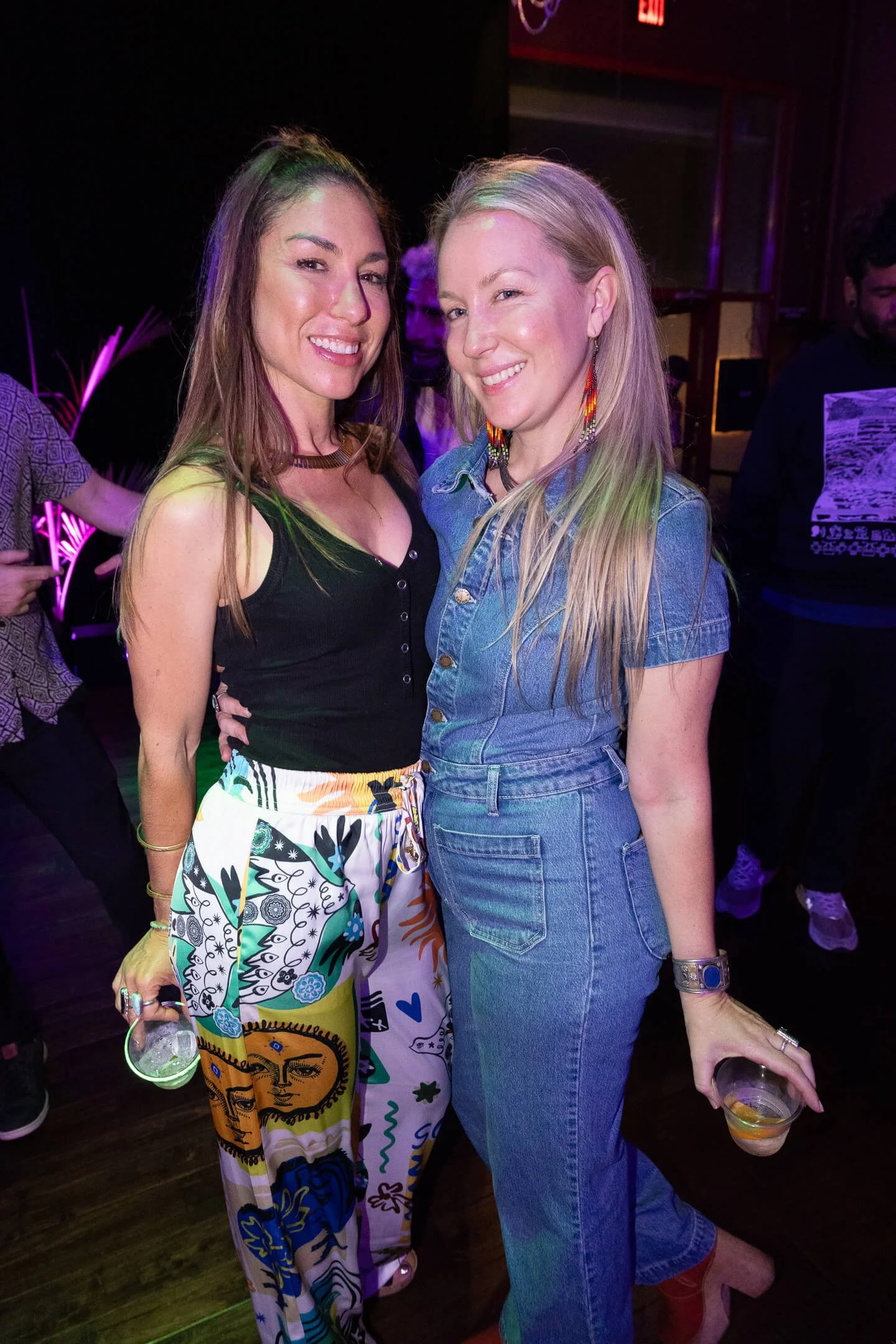 Two women standing close together at a party, smiling at the camera with drinks in hand, under vibrant purple lighting.