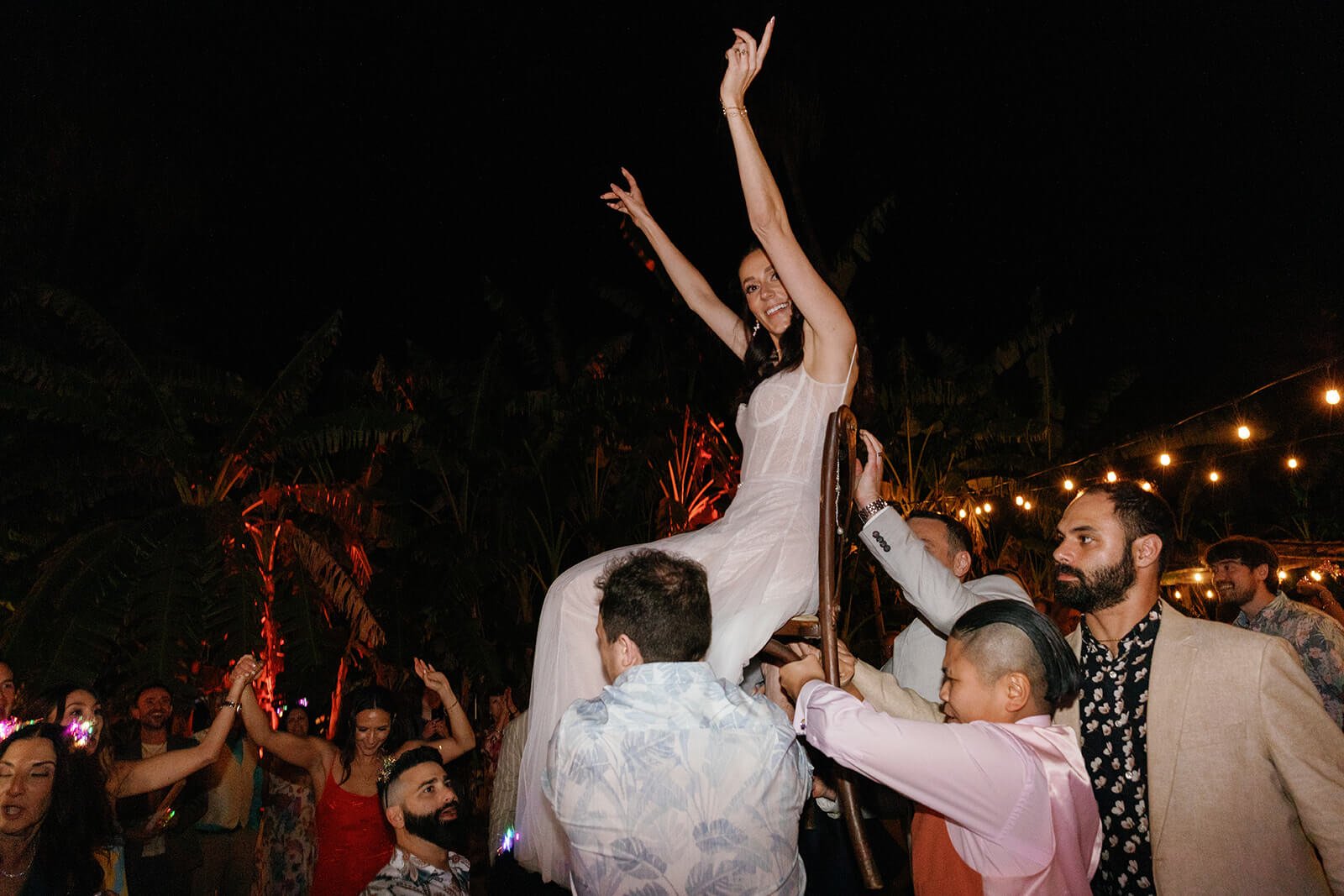 A woman in a white dress sitting on a chair being lifted by multiple men at an outdoor night celebration with string lights.