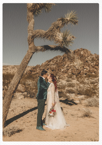 A couple in wedding attire kissing under a tall, crooked desert tree with a rocky background, in a desert landscape.