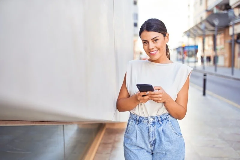 A young woman with dark hair in a ponytail, wearing a white sleeveless top and blue jeans, smiling and looking at her smartphone while walking outdoors on a city street.