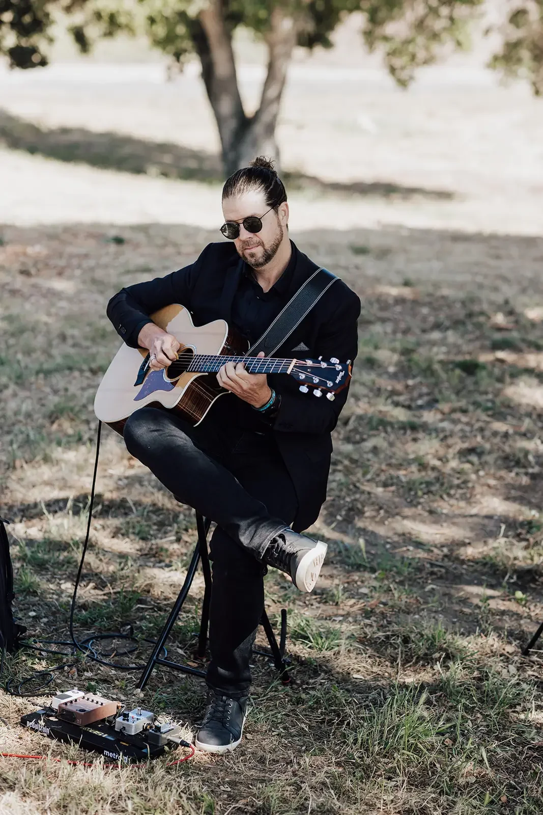 A man with long hair tied back, wearing sunglasses and a black jacket, is sitting outdoors on a small stool playing an acoustic guitar. There are guitar pedals on the ground near him, and he is in front of a tree with a grassy, slightly sandy area underneath.