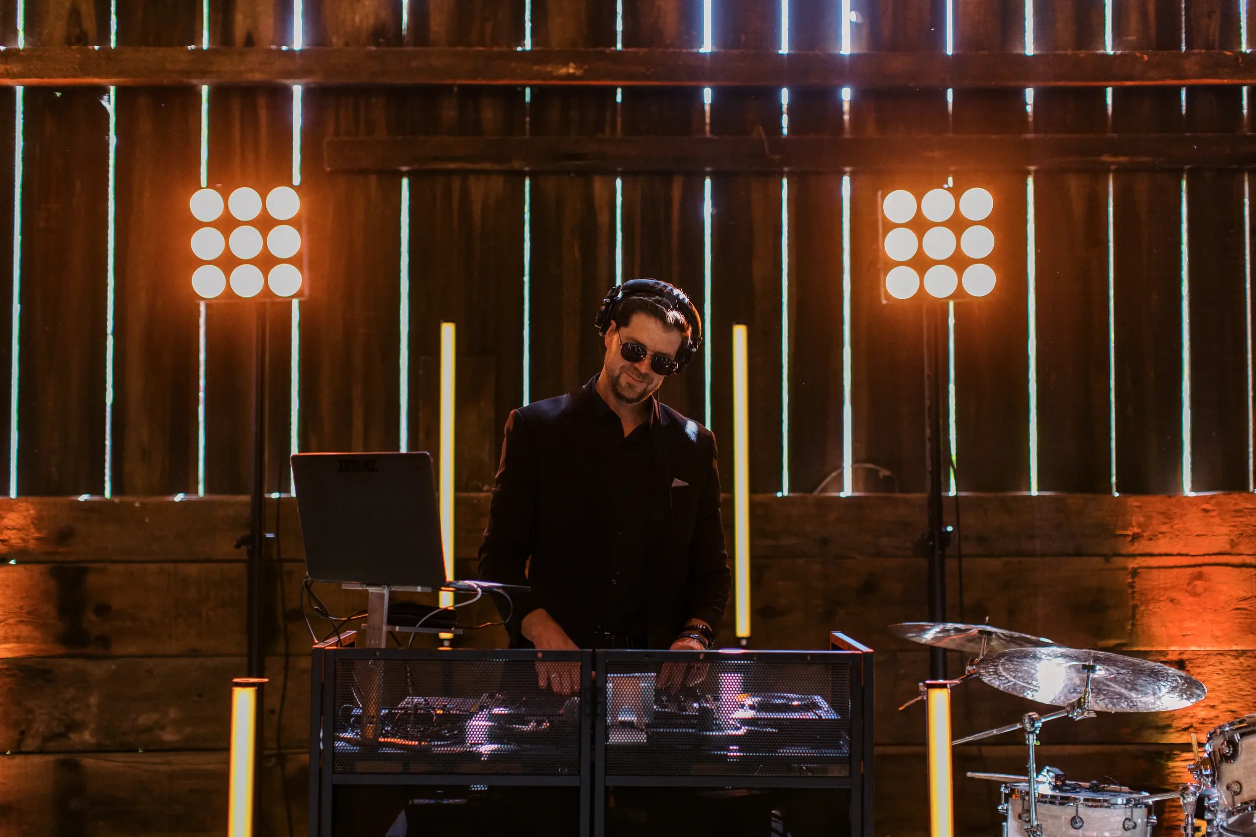 DJ in black clothing and sunglasses wearing headphones, standing behind DJ equipment and mixing music in a dimly lit space with colorful lights and wooden backdrop.