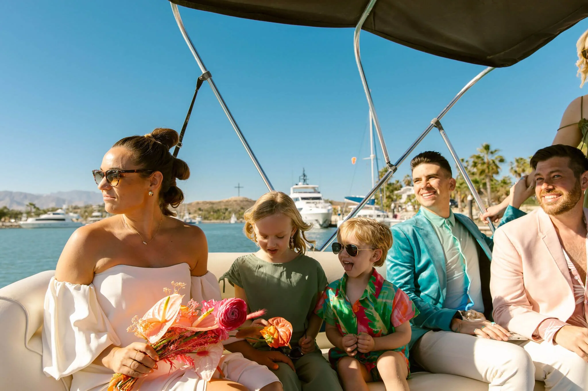 A family celebrating on a boat, with a woman holding a bouquet of flowers, children, and men dressed in bright suits, with boats and a cross visible in the background under a clear blue sky.