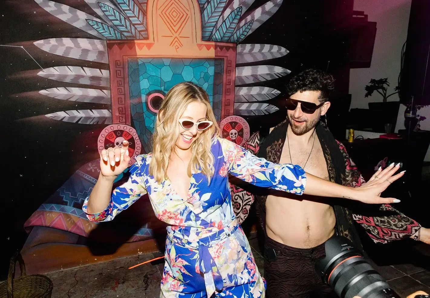 Two people dancing and smiling in front of a colorful, tribal-inspired backdrop with geometric patterns, feathers, and a skull motif. They are wearing sunglasses and festival attire.