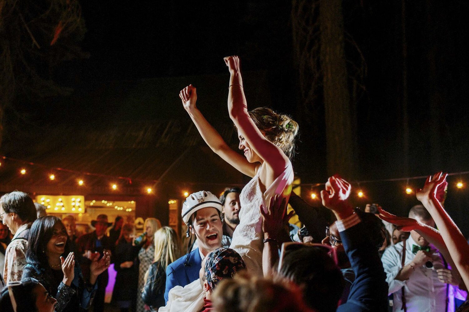 A woman is lifted on a person's shoulders at a celebration or party, with other people dancing and laughing around her in a festive setting with string lights and a decorated background.