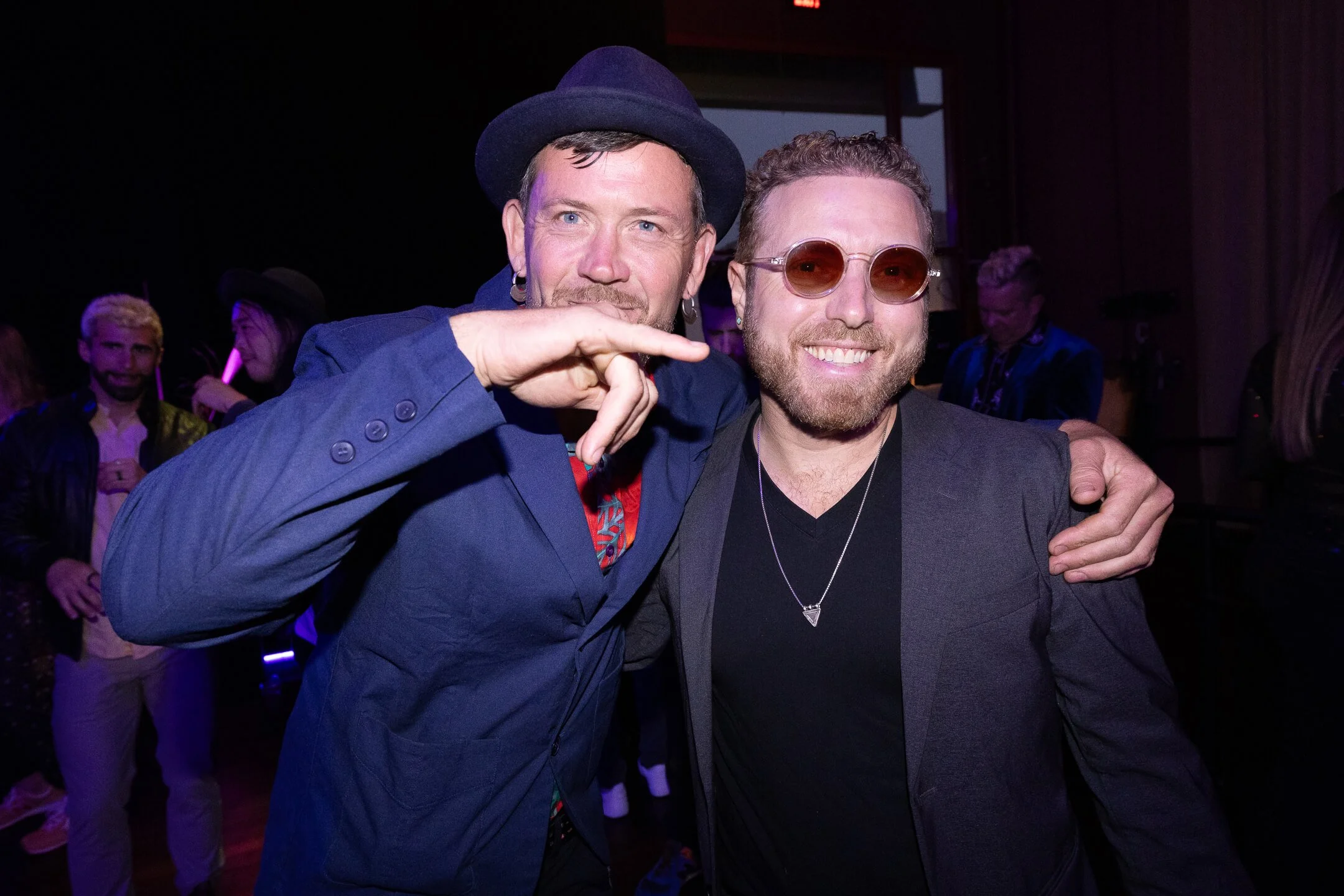 Two men smiling and posing for a photo at a party or event, with other people in the background.