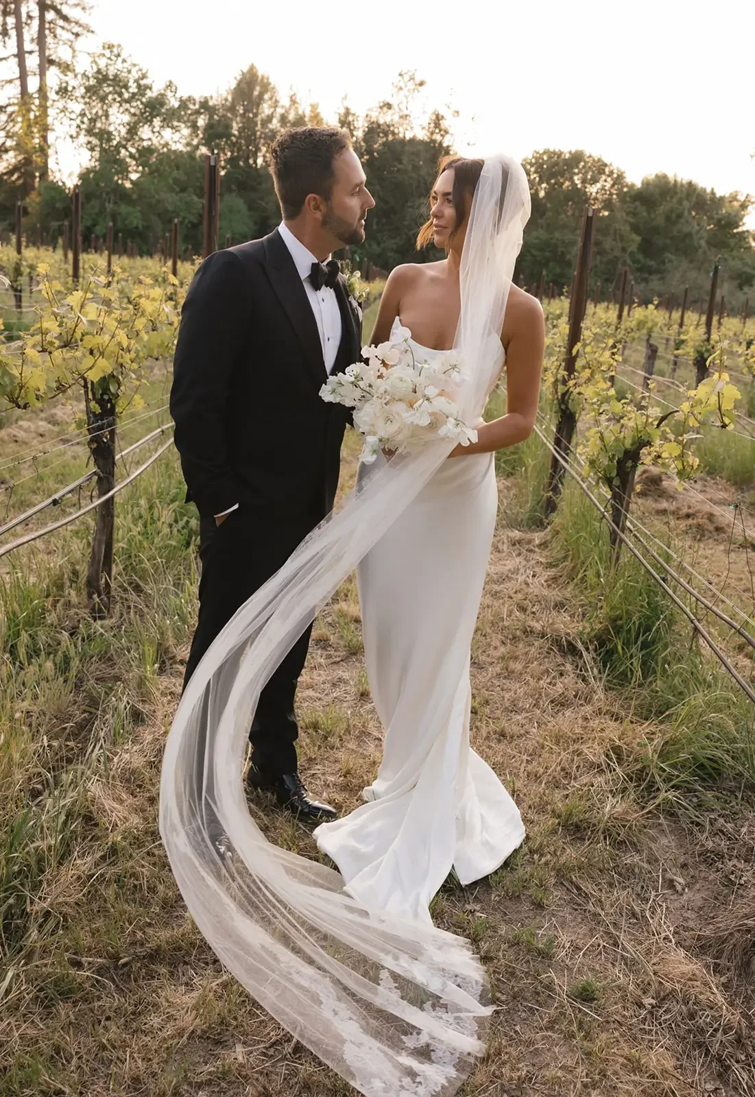 A bride and groom standing in a vineyard during sunset, facing each other. The bride is holding a bouquet of white flowers and wearing a white gown with a long veil. The groom is dressed in a black tuxedo with a bow tie.