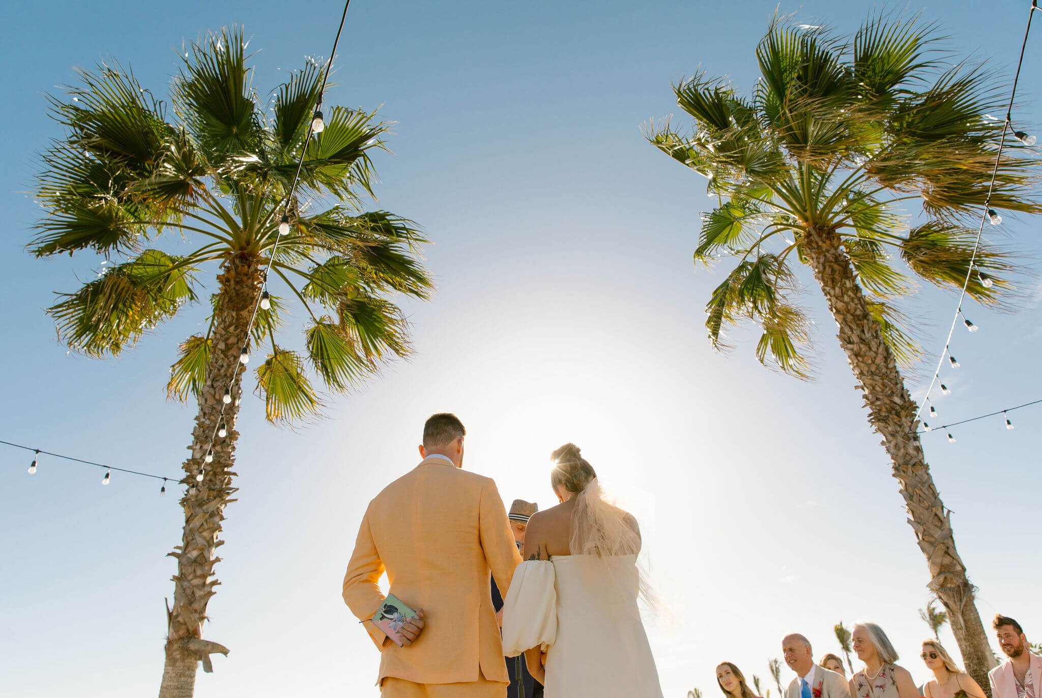 A wedding ceremony outdoors with two palm trees and string lights, sunlight shining behind the couple, with wedding guests in the background.