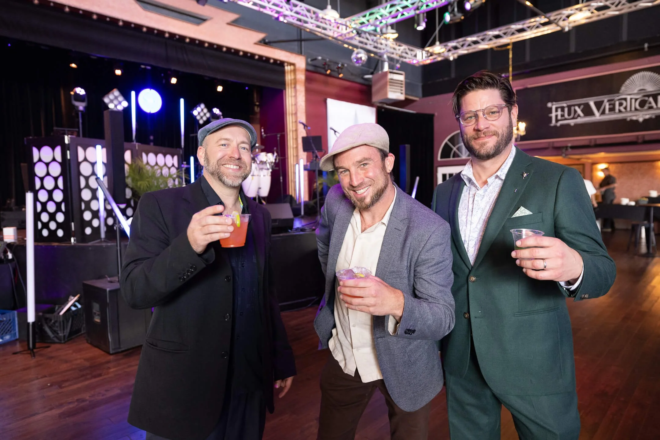 Three men in suits smiling and holding drinks at a party or social event in a venue with stage lights and a sign that reads 'Flux Vercain'.