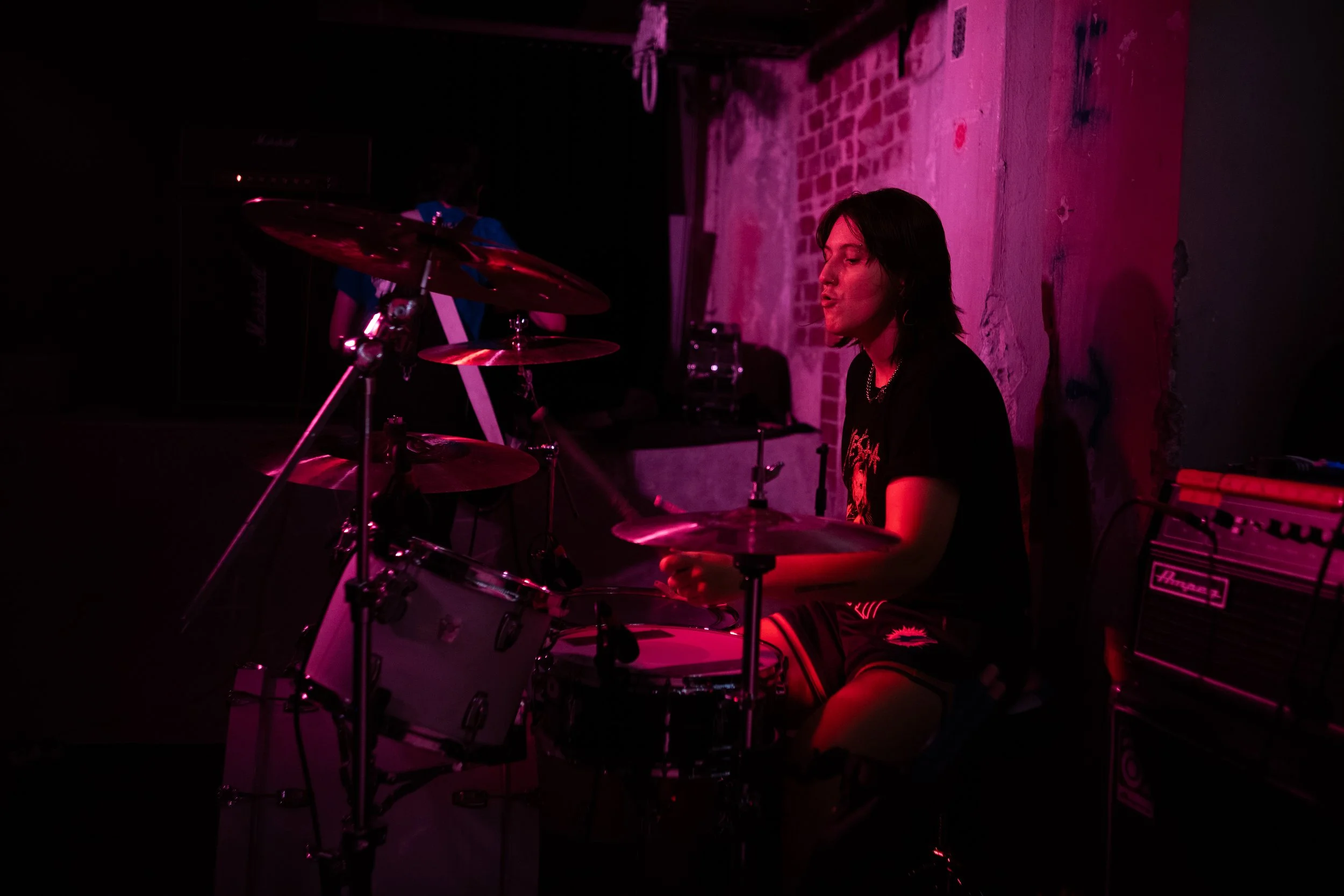 A woman playing drums in a dimly lit room with pink/purple lighting, brick wall background, and music equipment nearby.