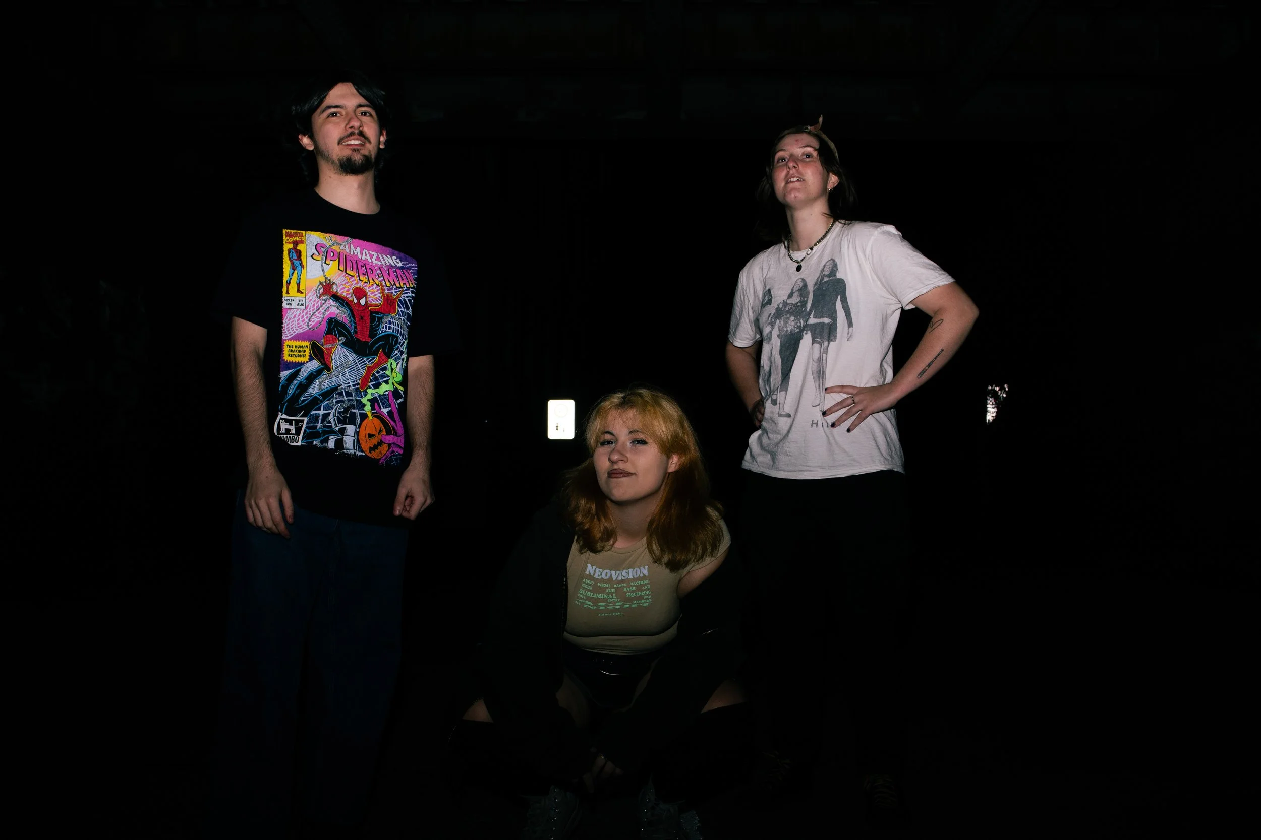 Three young adults in a dark setting, one sitting and two standing, with minimal lighting highlighting their upper bodies.
