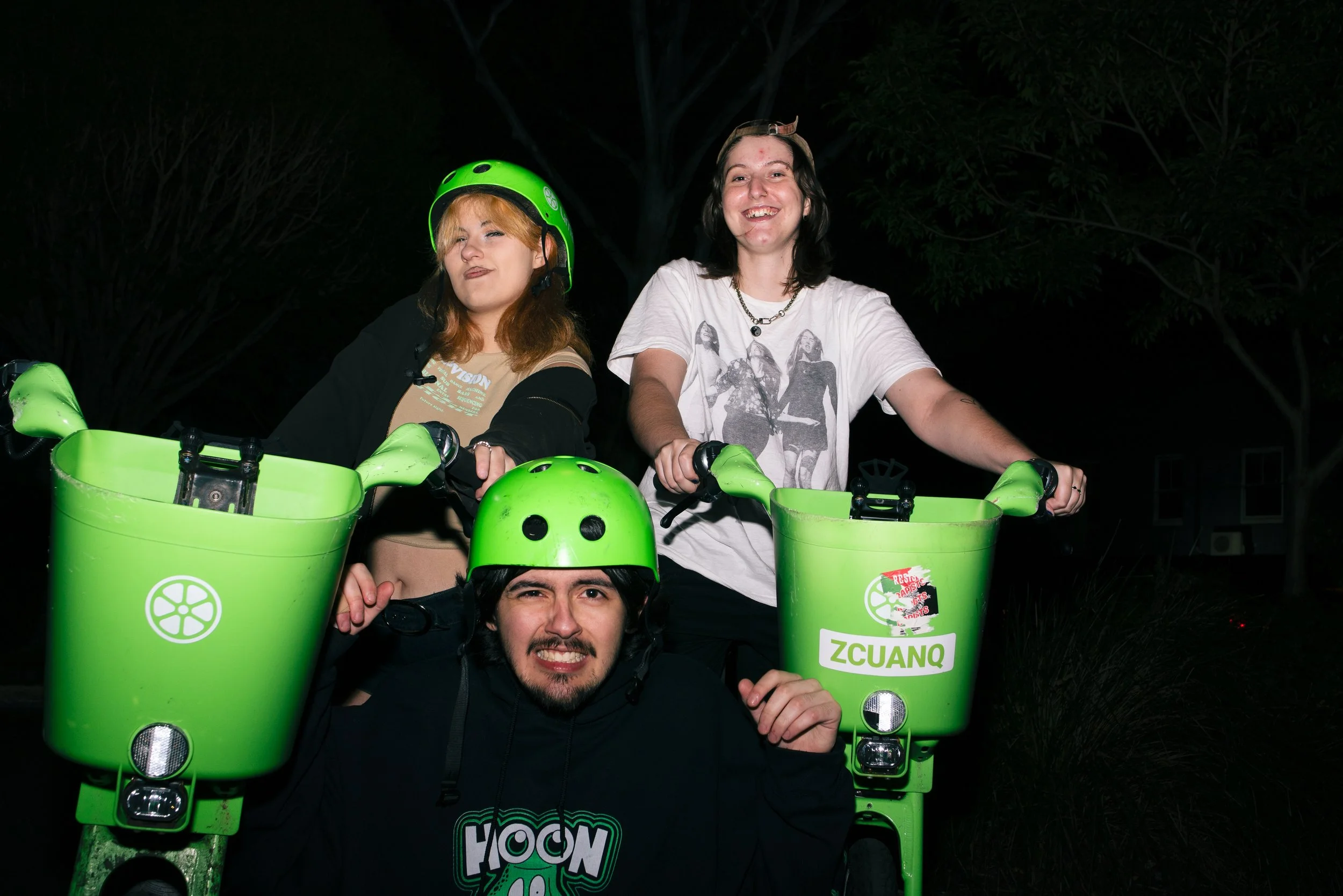 Three people with green delivery bikes and helmets smiling at night, two women standing behind and a man crouched in front, all posing outdoors.