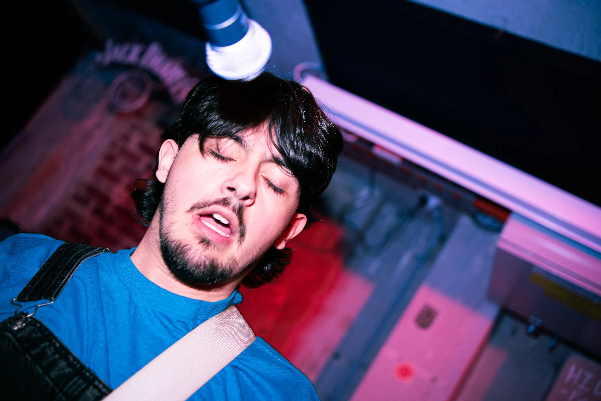 A man with black hair, facial hair, and a blue shirt, appears to be singing or performing with his eyes closed, in a setting with colorful lighting, a microphone below him, and a brick wall in the background.