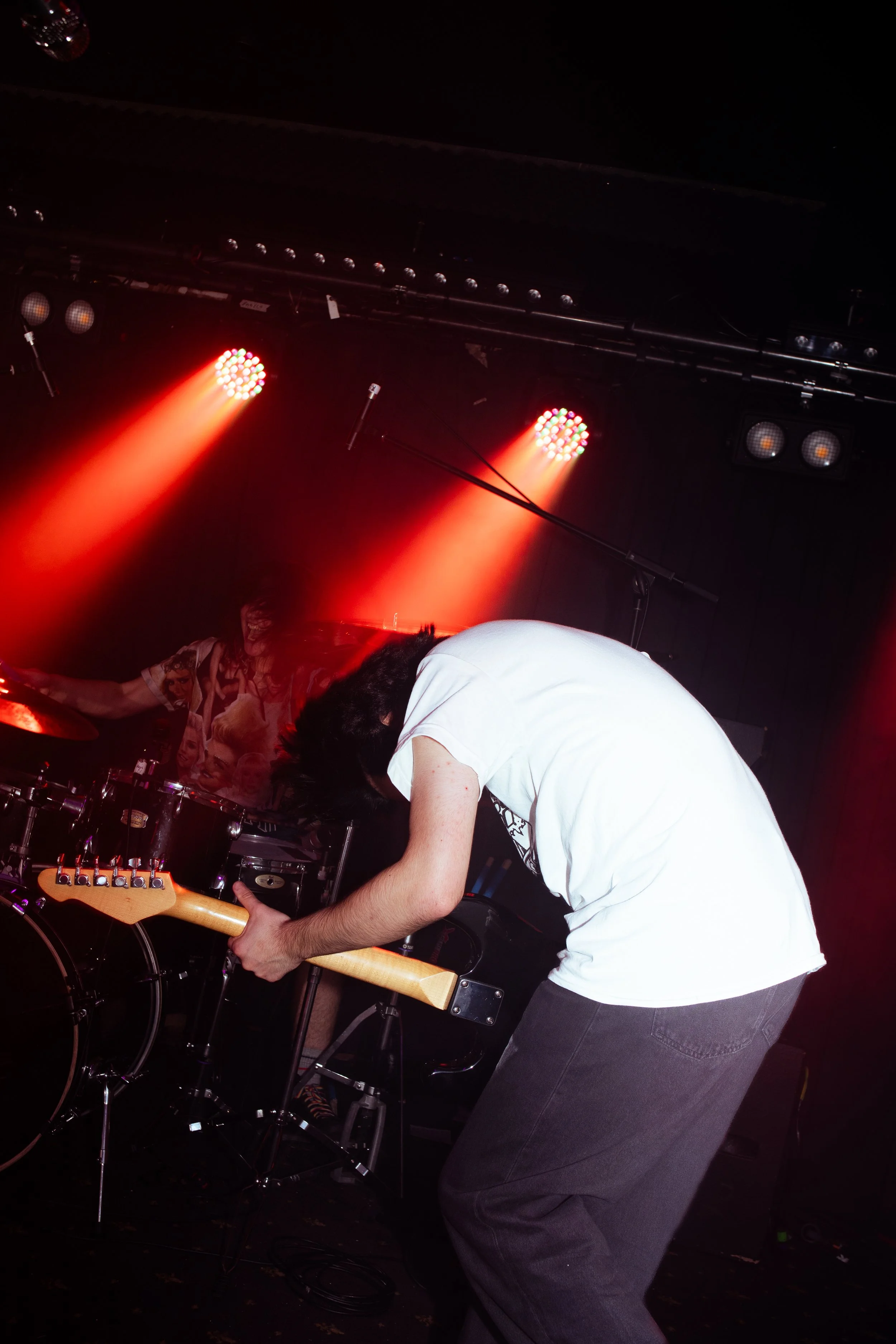 A musician playing an electric guitar on stage with red stage lights and a drummer in the background.