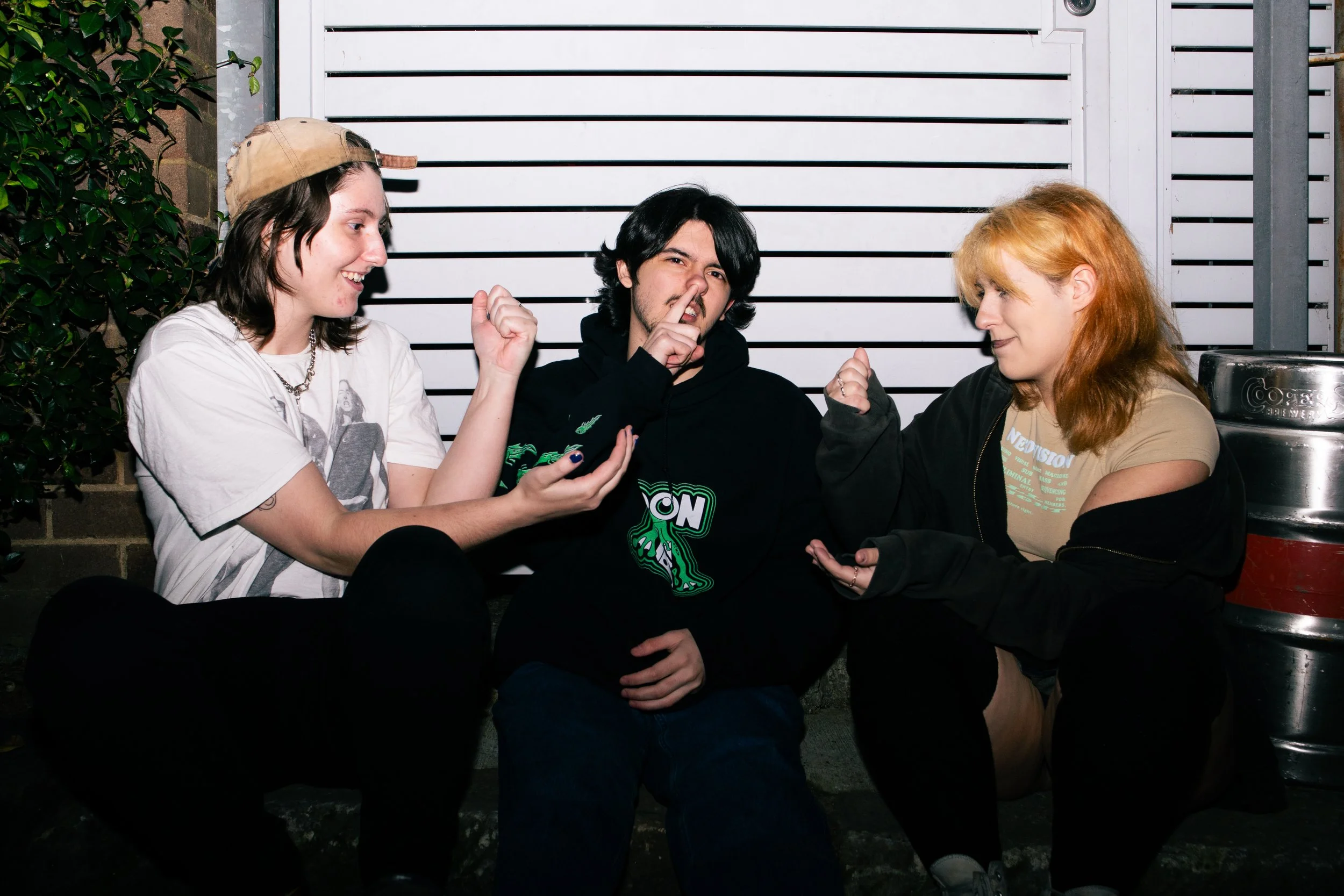 Three people sitting on pavement in front of a white fence, engaging in a playful interaction. The person on the left, wearing a beige cap backward, a white t-shirt, and dark pants, is smiling and gesturing with a fist. The person in the middle, wear