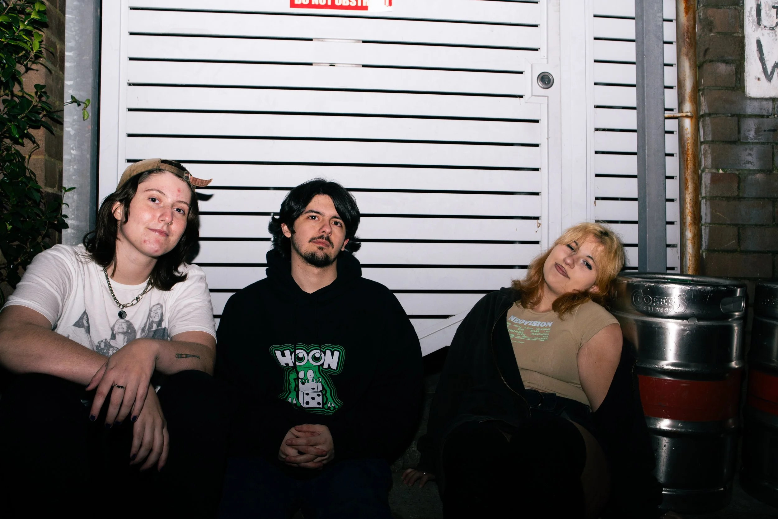 Three people sitting in front of a white metal gate. Two women on the left and right, and a man in the middle. The woman on the right is leaning against the metal barrels nearby. The setting appears to be outdoors at night.