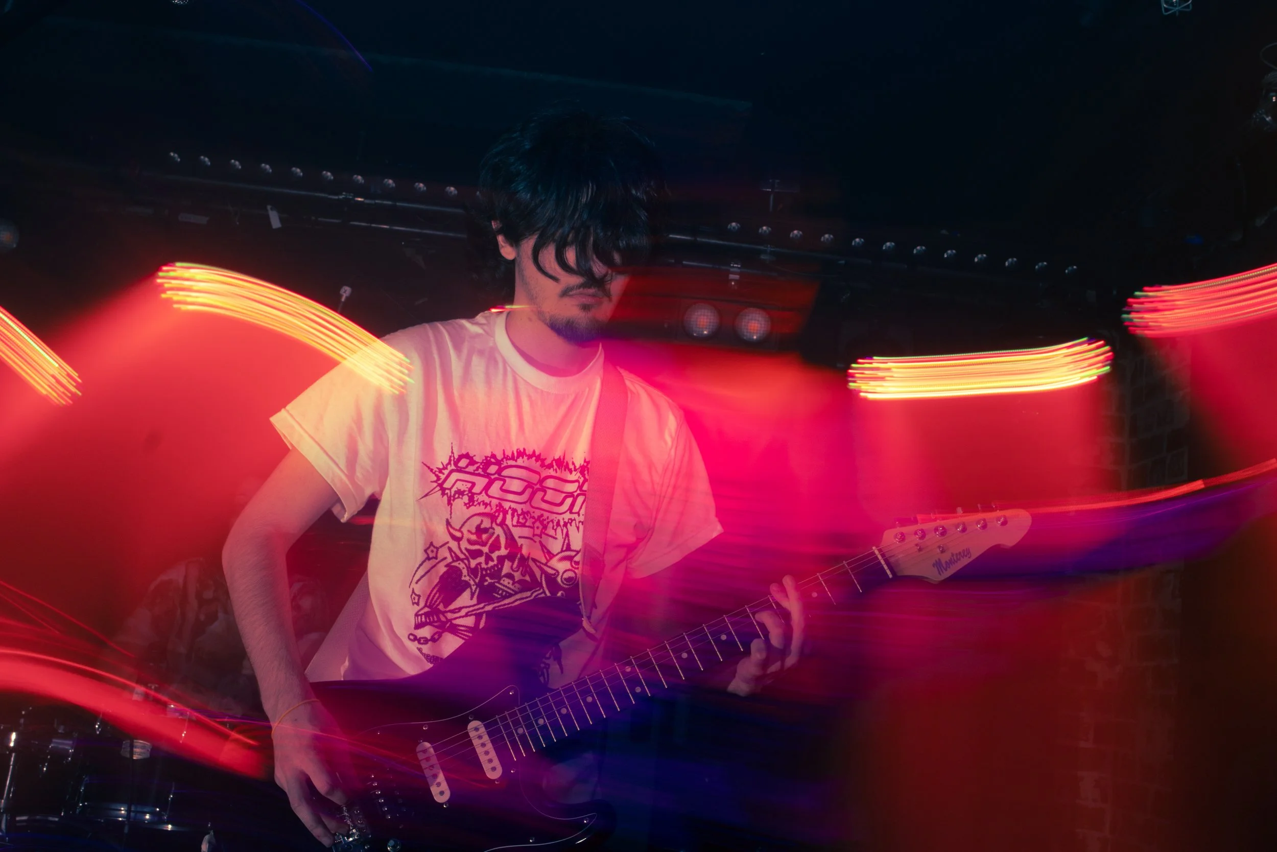 A musician plays an electric guitar on stage with vibrant red and pink light trails creating motion effects.