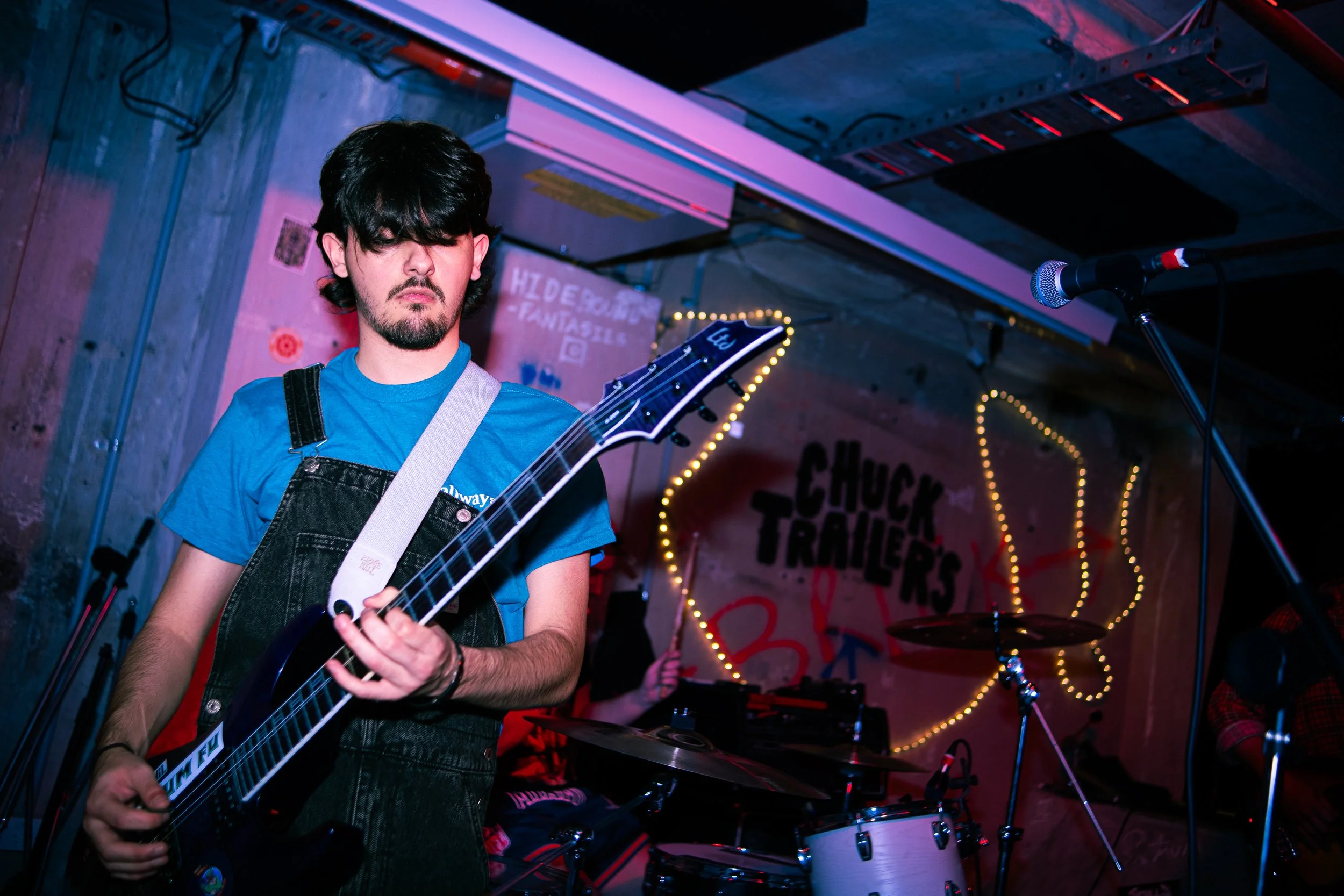 A young man with black hair and a goatee plays an electric guitar on a stage illuminated with colorful lights.