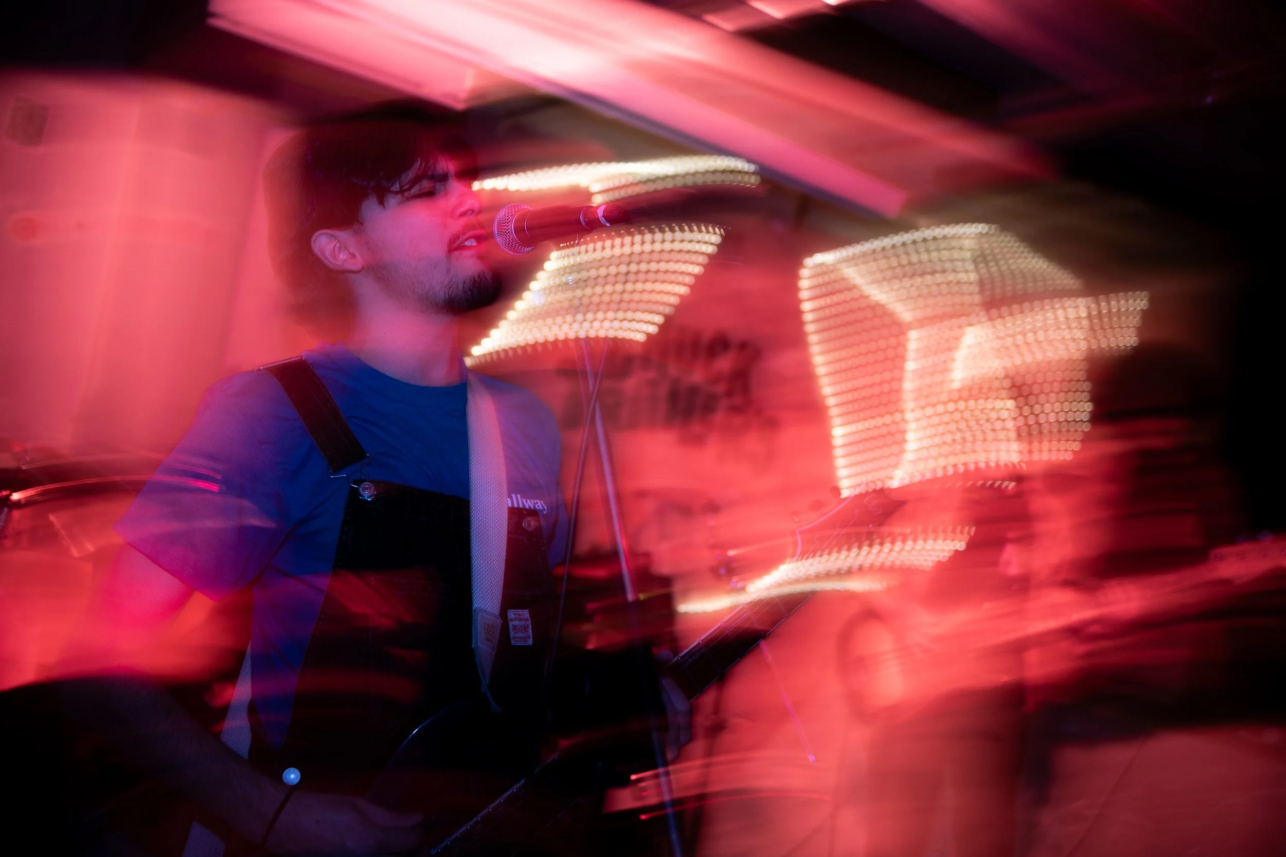 A man with dark hair and facial hair, wearing a blue shirt and black overalls, singing into a microphone while playing an electric guitar amidst colorful light streaks and blurred background.