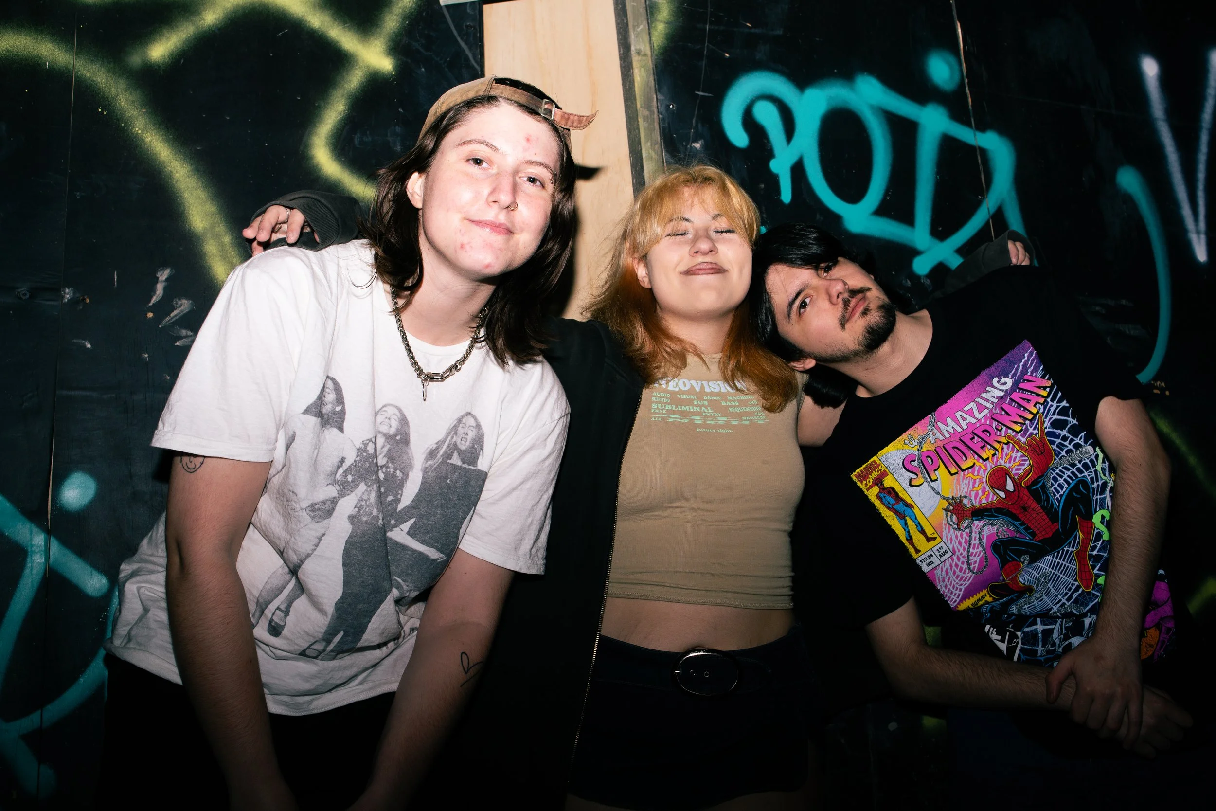 Three young adults standing close together in front of a graffiti-covered wall, smiling with eyes closed.