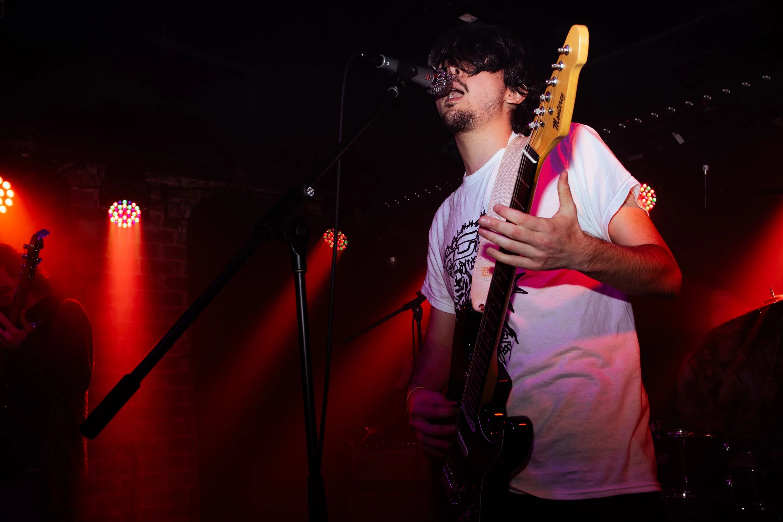 Male musician singing into a microphone while holding a black electric guitar on stage, with red and orange stage lights illuminating the background.