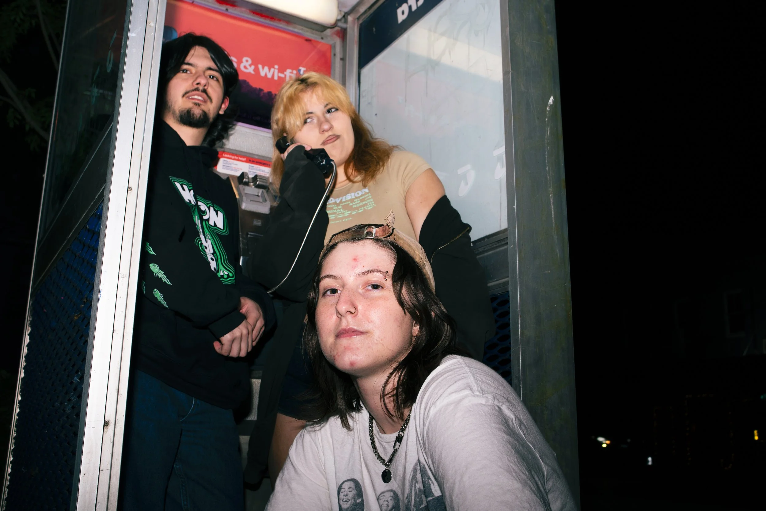 Three young people inside a phone booth at night, one with dark hair and a beard talking on the phone, another with blonde hair looking to the side, and a third with long dark hair and a cap sitting in front, wearing a graphic t-shirt.