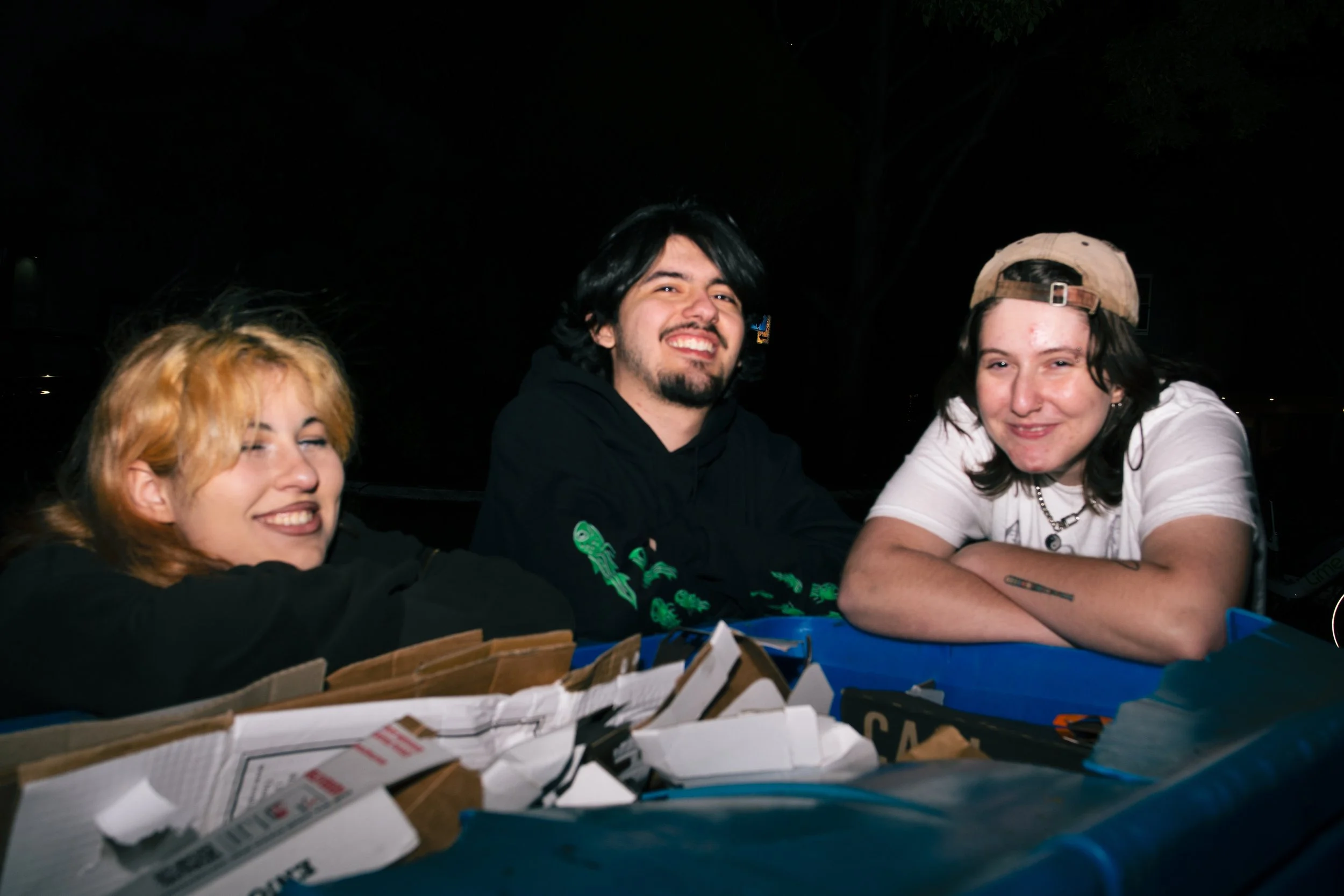 Three young adults sitting outdoors at night, smiling. Two women and one man, with cluttered cardboard boxes and containers in front of them.