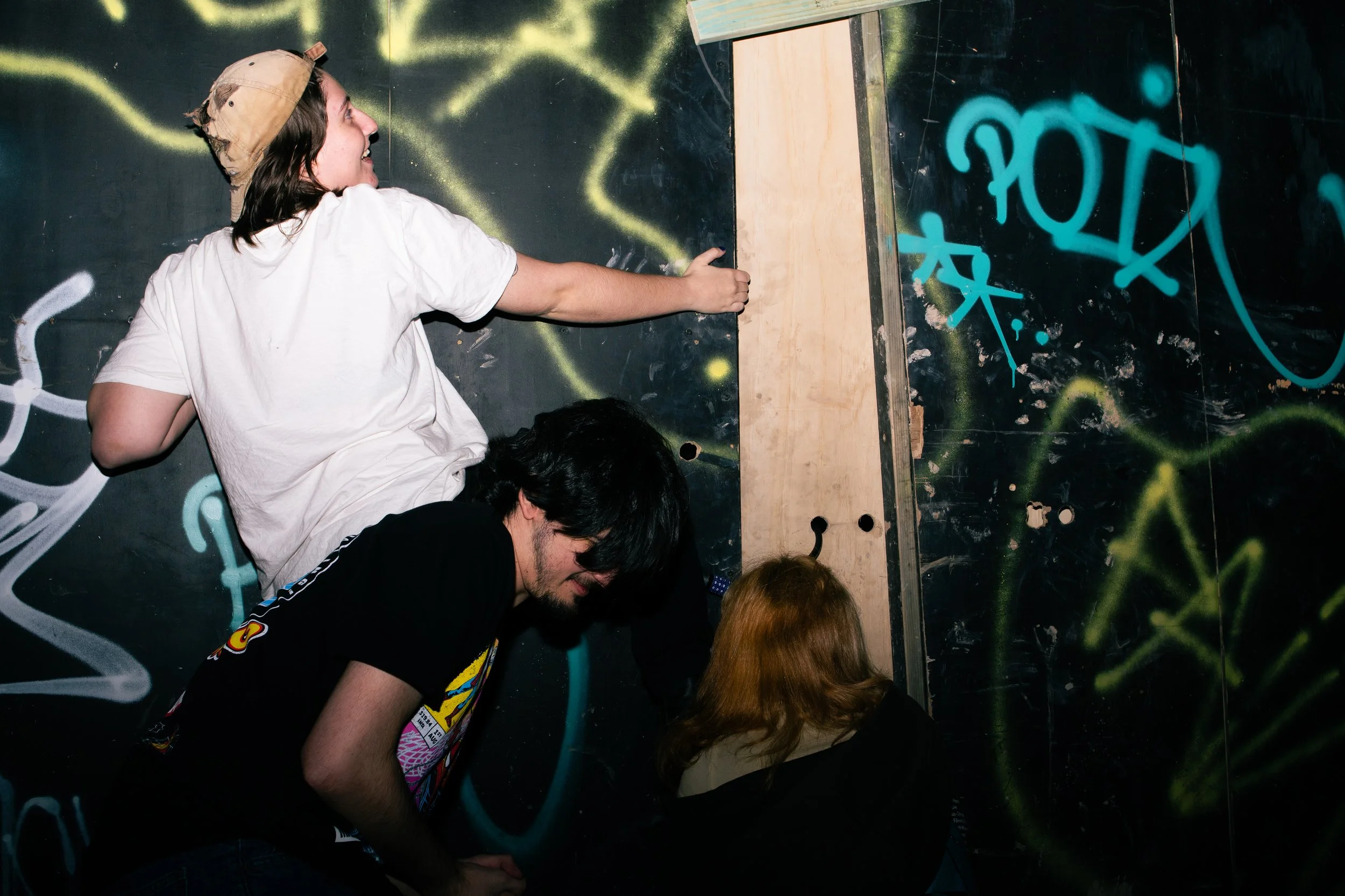 Four people in an indoor graffiti-covered space with a black wall, one of whom is climbing a makeshift wooden wall while the others watch.
