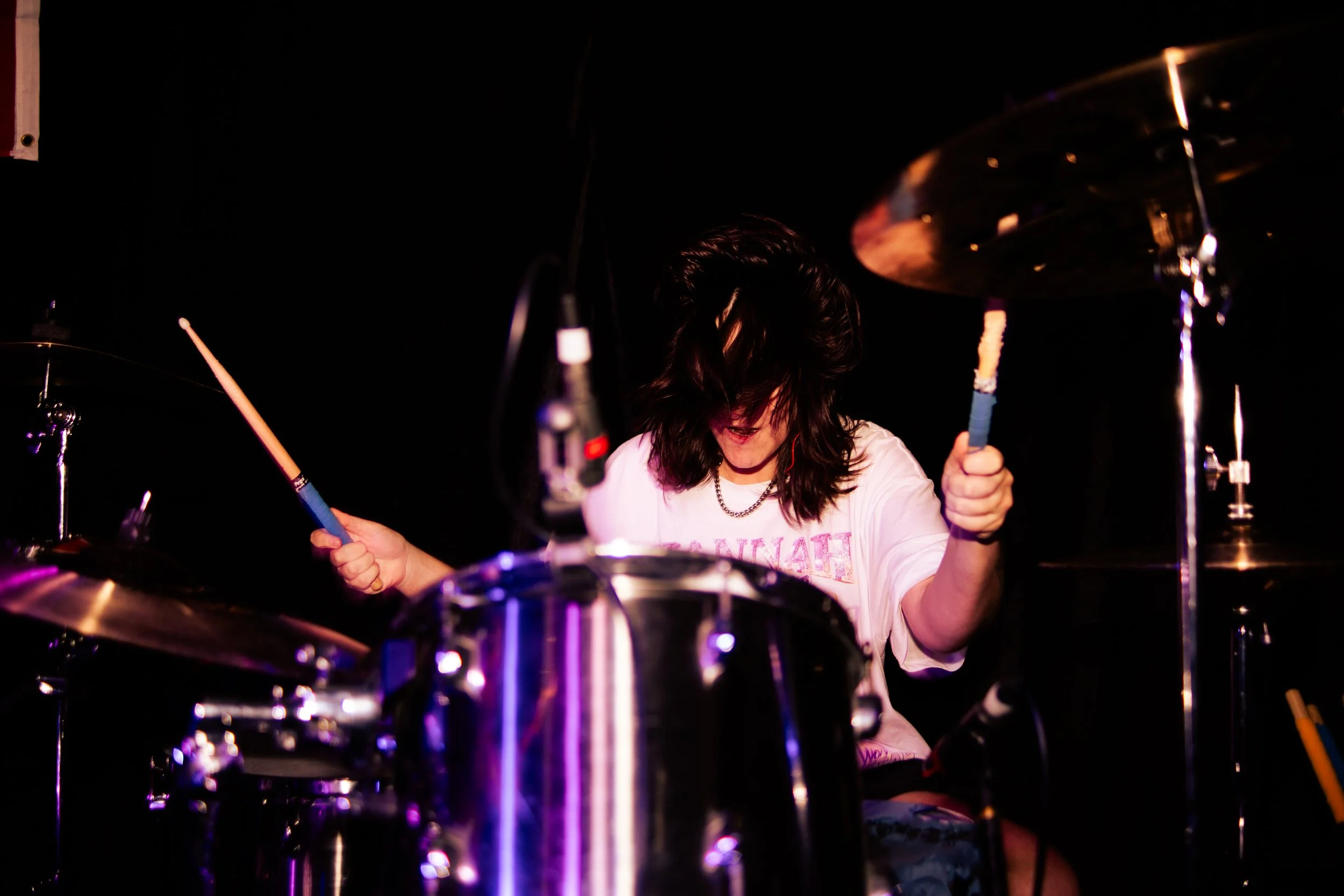 Person with long dark hair playing drums in a dark setting, wearing a white t-shirt and holding drumsticks.